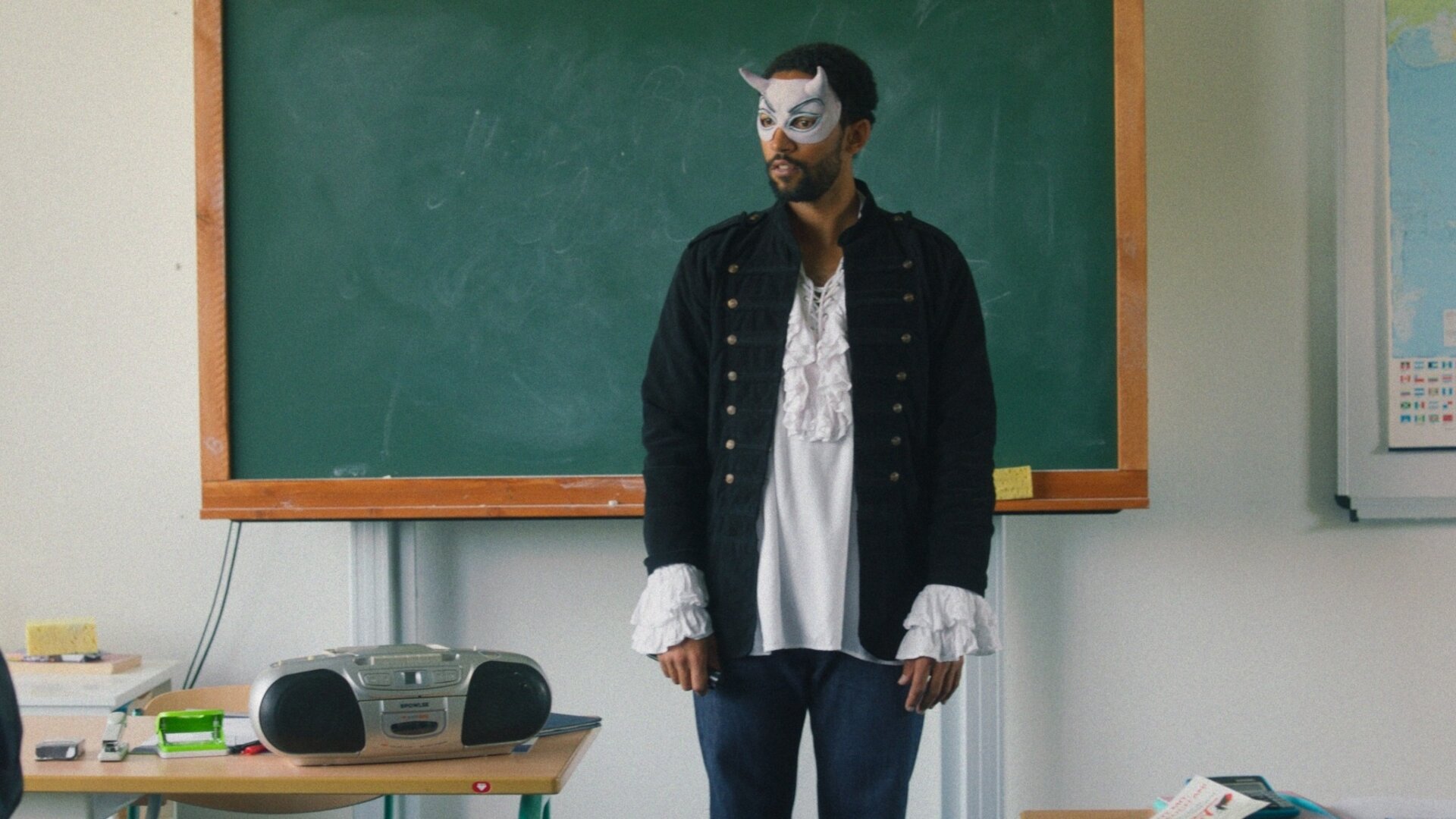 "Doppelhaushälfte – Musical": Theo Kröger (Benito Bause) steht verkleidet als Mephisto vor einer Tafel in einem Klassenzimmer. Er trägt eine Maske und historisches Kostüm, vor ihm liegen Unterrichtsmaterialien.