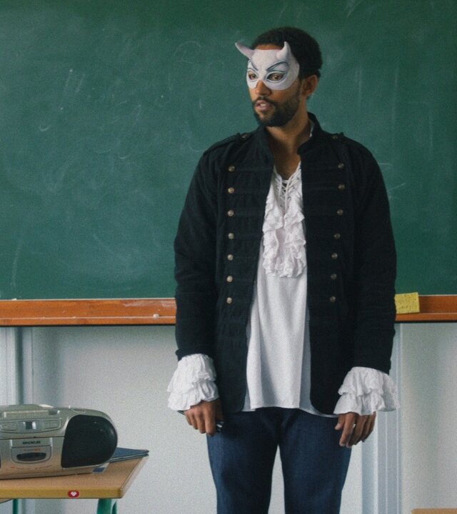 "Doppelhaushälfte – Musical": Theo Kröger (Benito Bause) steht verkleidet als Mephisto vor einer Tafel in einem Klassenzimmer. Er trägt eine Maske und historisches Kostüm, vor ihm liegen Unterrichtsmaterialien.