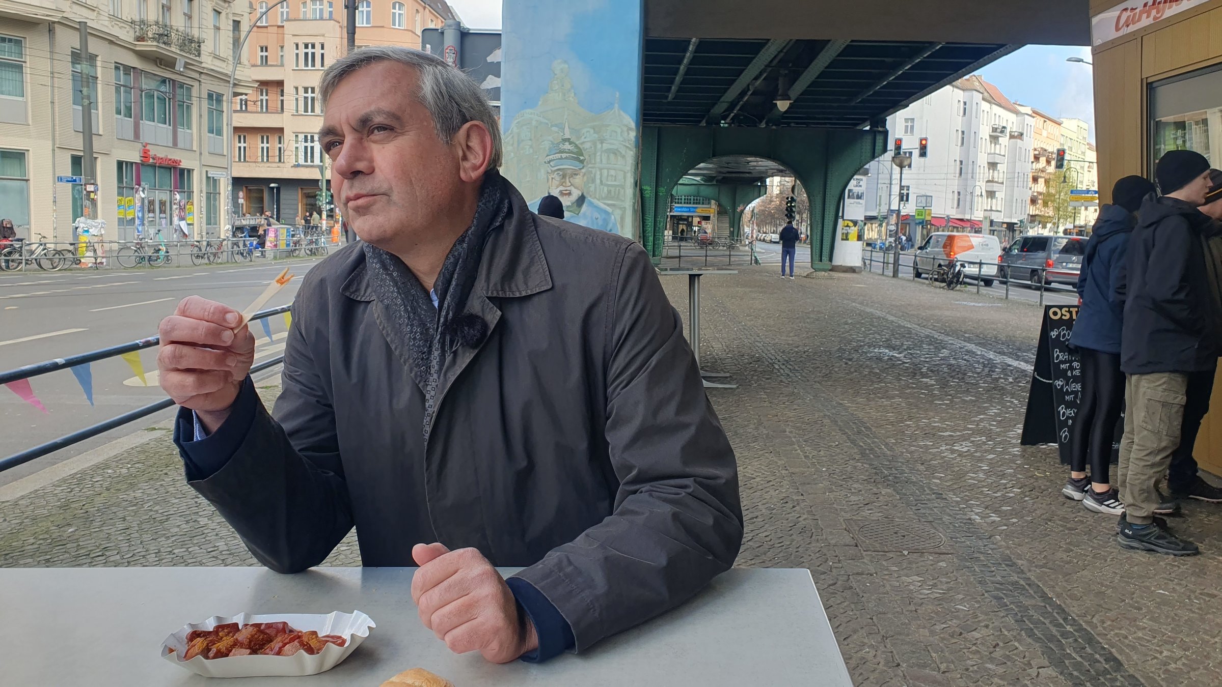 "Kaminer Inside: Wie isst Deutschland?": Kaminer steht unter einem U-Bahn-Bogen an einem Stehtisch und verspeist genüsslich eine Currywurst. Am Torbogen hinter ihm als Graffiti ein preußischer Offizier mit Pickelhaube.