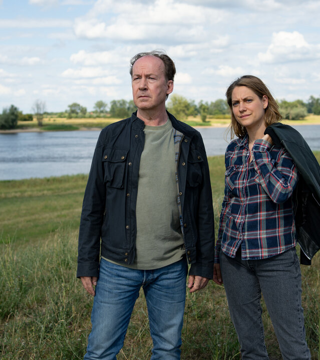 "Wendland - Stiller und der rote Faden": Jakob Stiller (Ulrich Noethen) und Kira Engelmann (Bettina Burchard) stehen zusammen auf einer Wiese am Ufer.