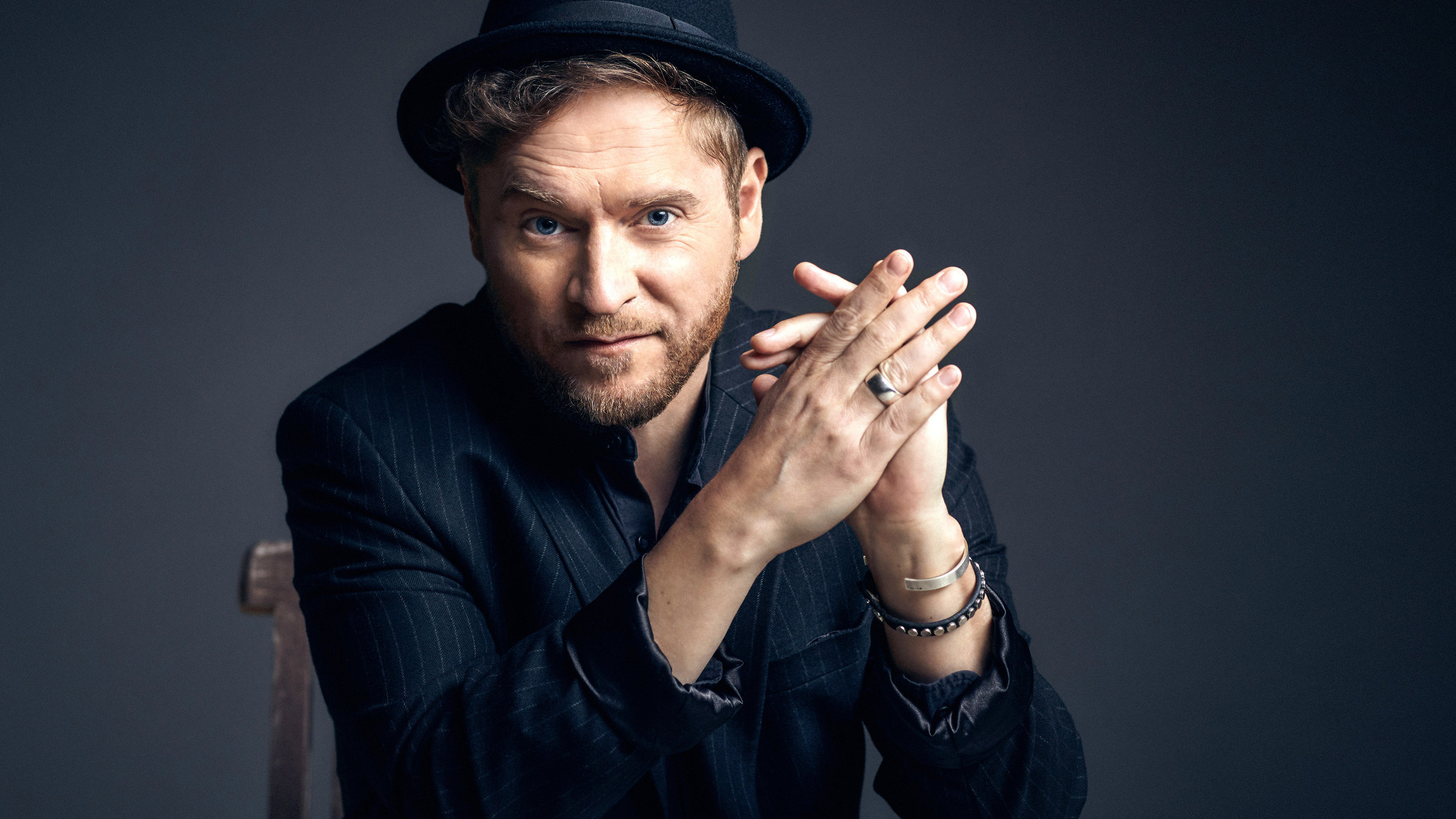 "Johannes Oerding: Live am Kalkberg": Portrait-Foto des Sängers Johannes Oerding. Er sitzt vor einer dunklen Wand und blickt in die Kamera. Die Hände sind seitlich vor dem Gesicht gefaltet. Bekleidet ist er mit einem dunklen Nadelstreifen-Jacket und einem schwarzen Hut.