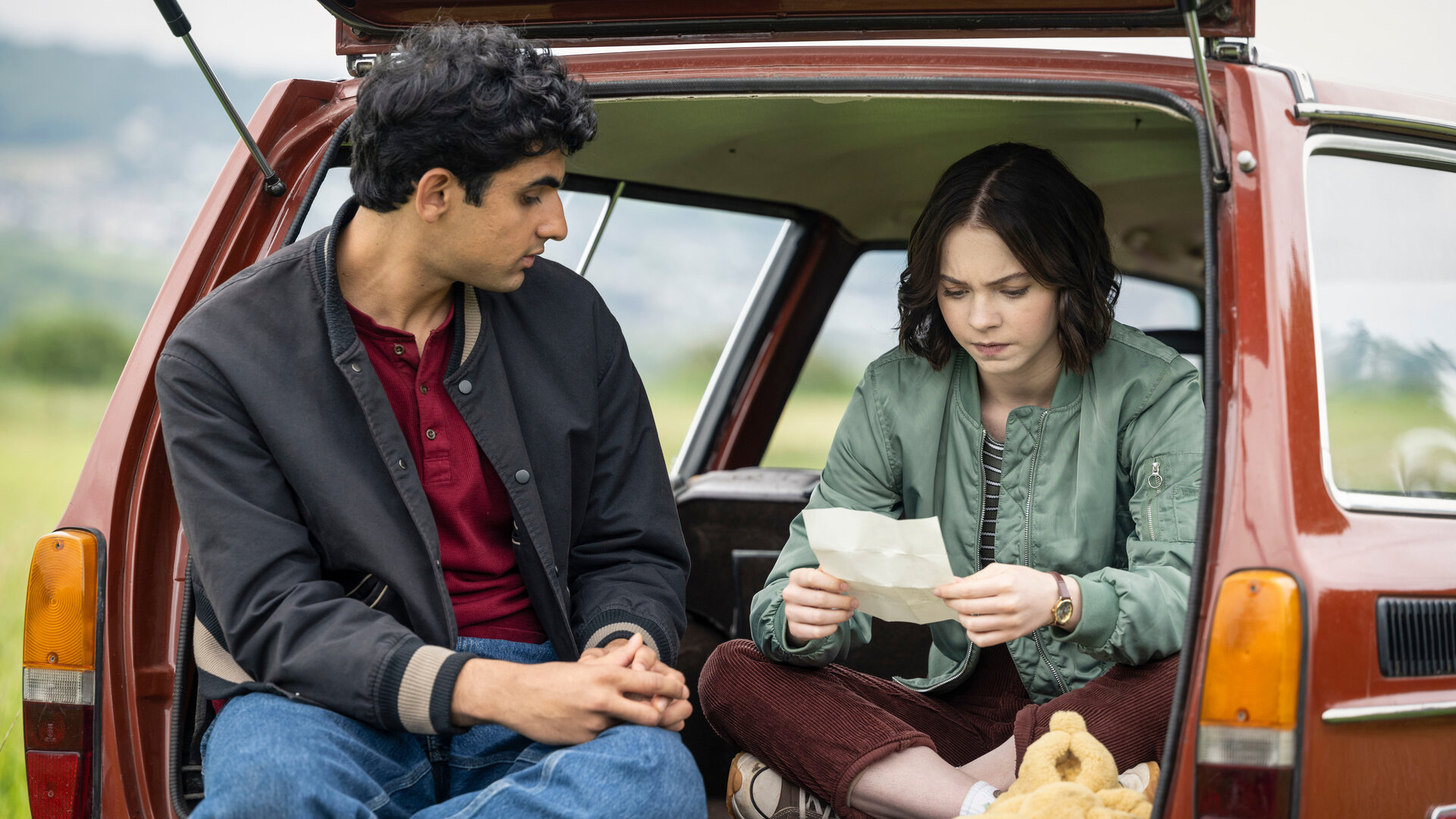 „A Good Girl's Guide to Murder – Wer war Andie Bell?“: Ravi (Zain Iqbal) und Pip (Emma Myers) sitzen im geöffneten Kofferraum eines roten Autos, das in einem Feld parkt. Pip studiert mit ernstem Blick ein weißes, aufgefaltetes Blatt, während Ravi sie von links daneben betrachtet. Vor Pip liegt zudem ein gelbes Kuscheltier.