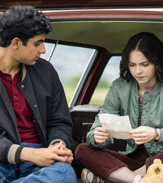 „A Good Girl's Guide to Murder – Wer war Andie Bell?“: Ravi (Zain Iqbal) und Pip (Emma Myers) sitzen im geöffneten Kofferraum eines roten Autos, das in einem Feld parkt. Pip studiert mit ernstem Blick ein weißes, aufgefaltetes Blatt, während Ravi sie von links daneben betrachtet. Vor Pip liegt zudem ein gelbes Kuscheltier.