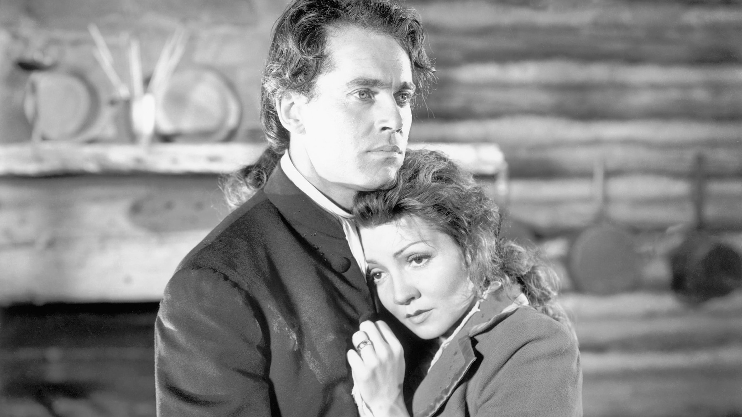 "Trommeln am Mohawk": Gilbert Martin (Henry Fonda) hält Lana (Claudette Colbert) in den Armen, beide schauen melancholisch. Im Hintergrund ist unscharf ein Teil des steinernen Kamins eines Blockhauses zu sehen.