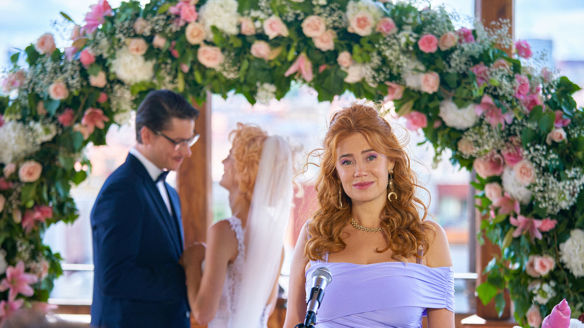 "My Ex - Die Liebenden": Jan (Šimon Obdržálek) und Emma (Alli Neumann) stehen unter einem begrünten Traubogen mit weißen und rosa Rosen und schauen sich sehr verliebt an. Ivy (Palina Rojinski) steht seitlich vor ihnen vor einem Mikrophon und hält eine Rede.