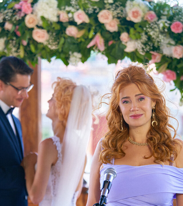 "My Ex - Die Liebenden": Jan (Šimon Obdržálek) und Emma (Alli Neumann) stehen unter einem begrünten Traubogen mit weißen und rosa Rosen und schauen sich sehr verliebt an. Ivy (Palina Rojinski) steht seitlich vor ihnen vor einem Mikrophon und hält eine Rede.