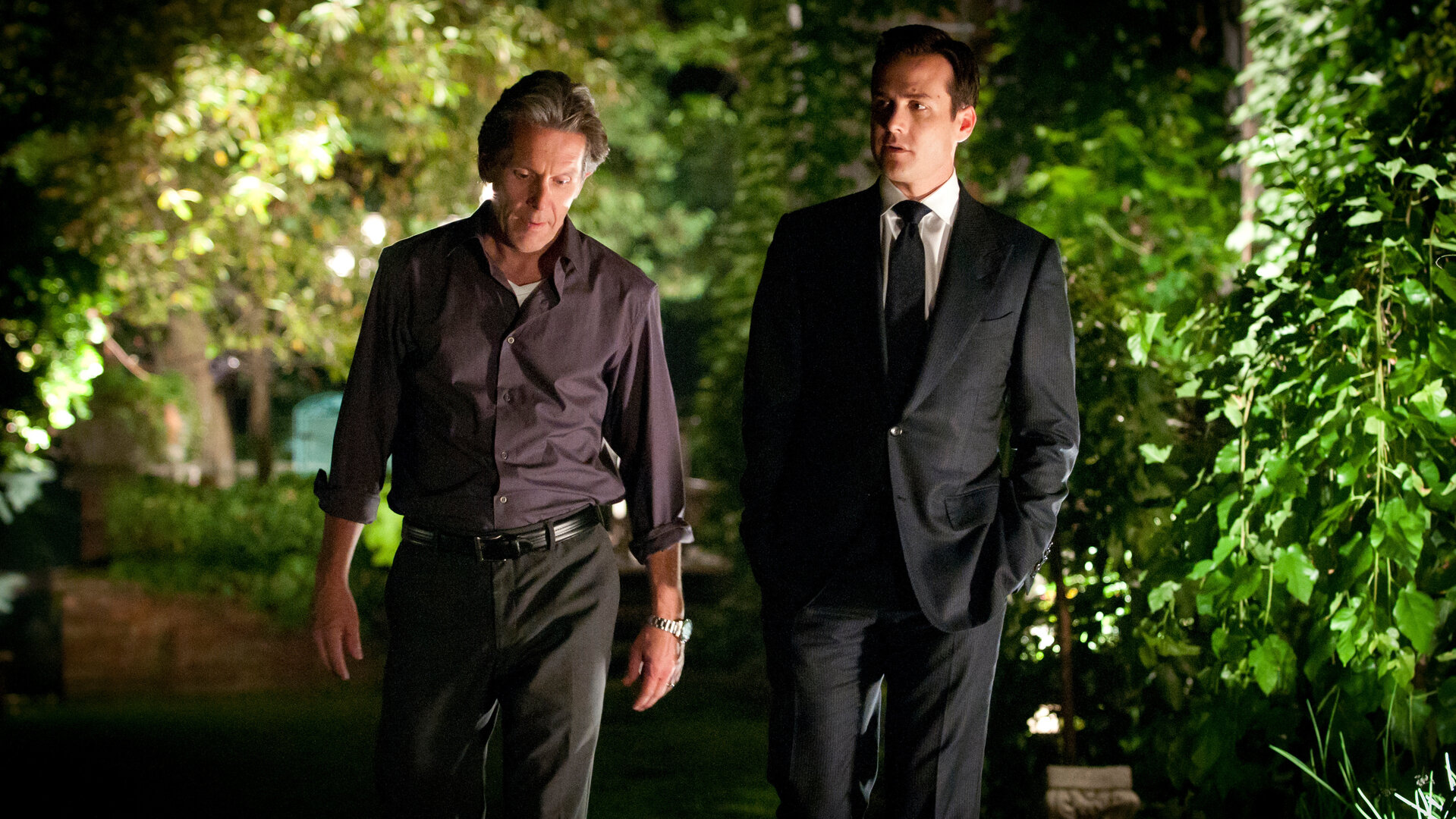 "Suits - Spielregeln": Harvey (Gabriel Macht) und Cameron Dennis (Gary Cole) gehen nebeneinander einen nächtlich beleuchteten Weg entlang. Während Harvey den Blick zur Seite richtet, schaut Cameron nach unten – beide wirken ernst.
