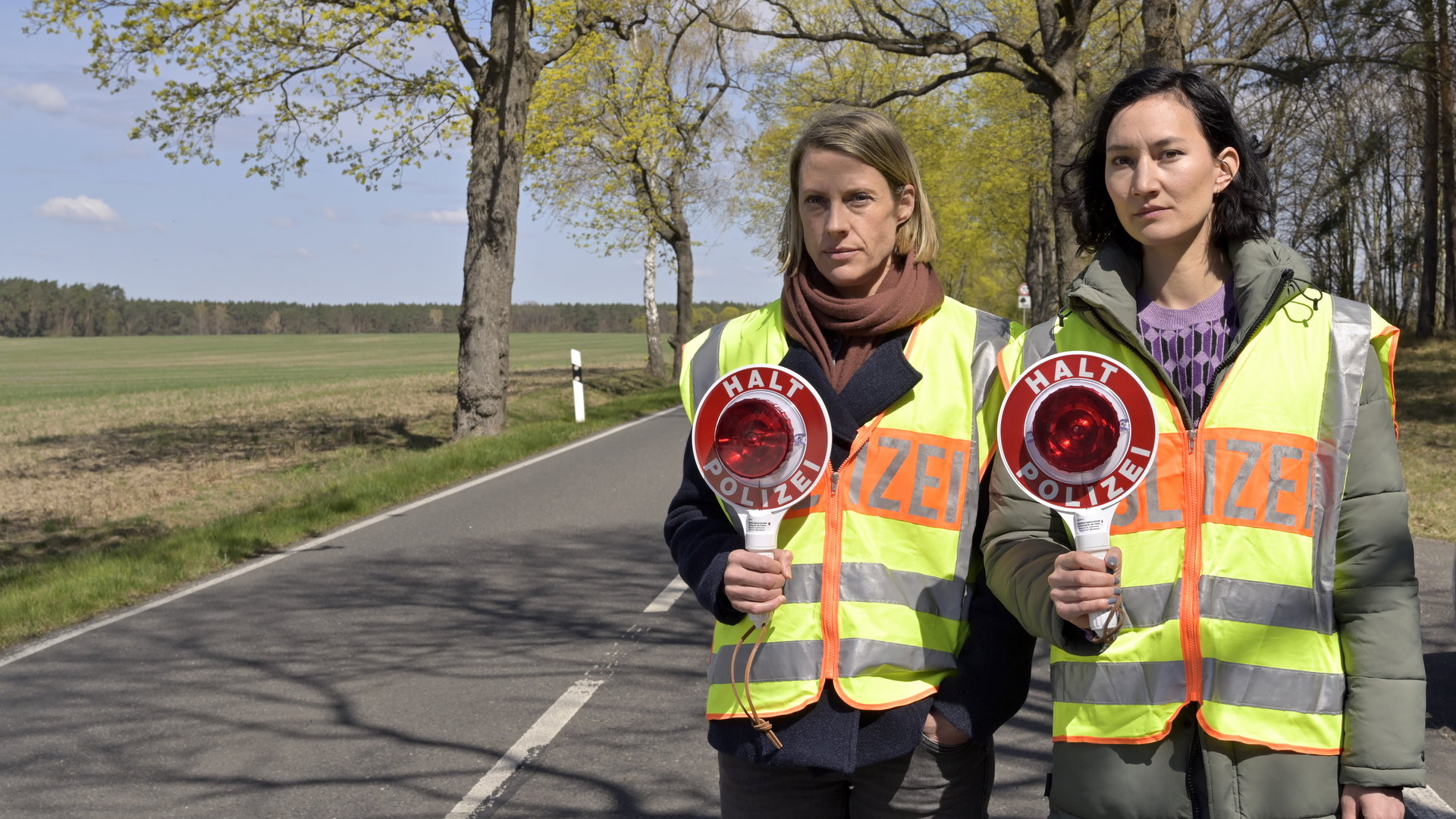 "SOKO Potsdam - Mensch und Maschine": Mitten auf einer Landstraße stehen Tamara Meurer (Anja Pahl) und Pauline Hobrecht (Agnes Decker). Beide tragen eine Polizei-Warnweste und halten eine Verkehrskelle in Richtung Kamera.