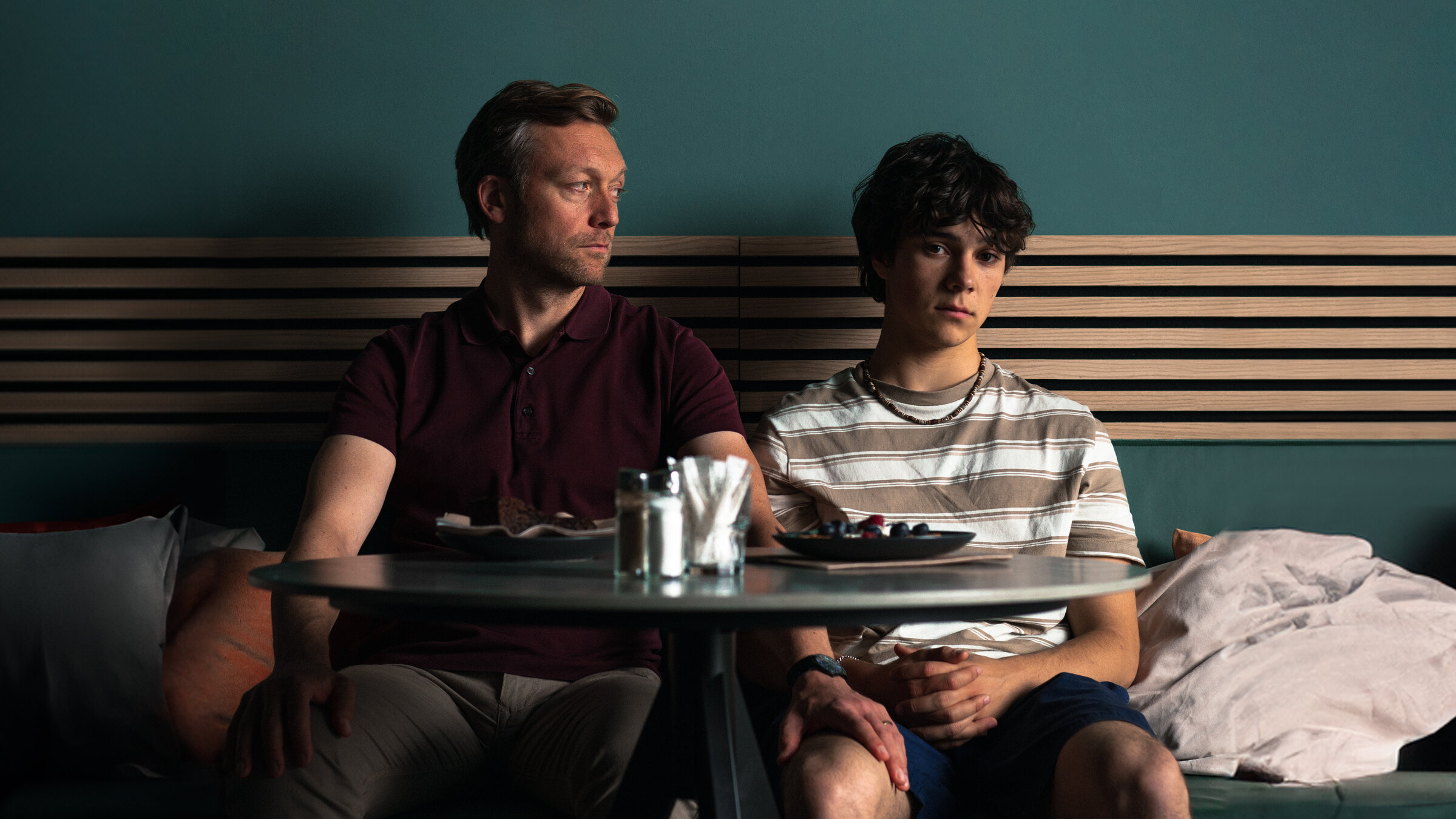 "No Dogs Allowed": Gabo (Carlo Krammling) und Dave (Robin Sondermann) sitzen nebeneinander an einem Cafétisch. Dave sieht zu Gabo und hat seine Hand auf dessen Oberschenkel gelegt. Gabo schaut gedankenverloren vor sich hin.