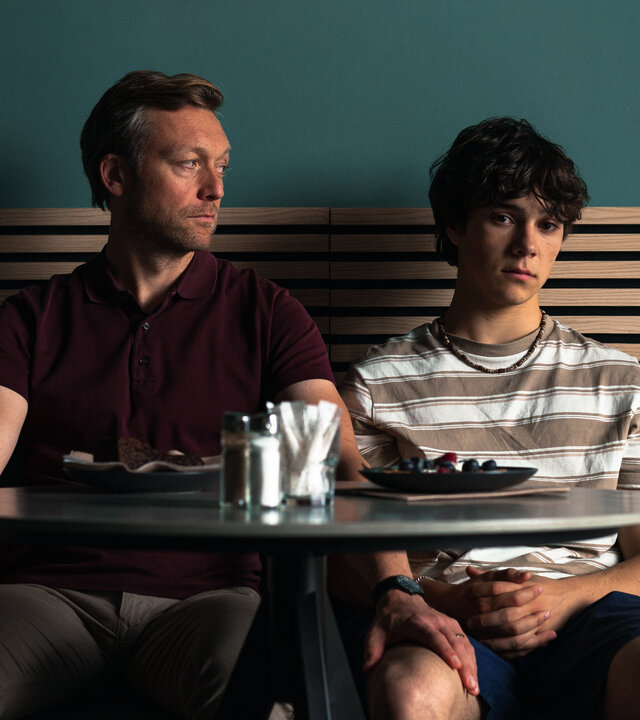 "No Dogs Allowed": Gabo (Carlo Krammling) und Dave (Robin Sondermann) sitzen nebeneinander an einem Cafétisch. Dave sieht zu Gabo und hat seine Hand auf dessen Oberschenkel gelegt. Gabo schaut gedankenverloren vor sich hin.