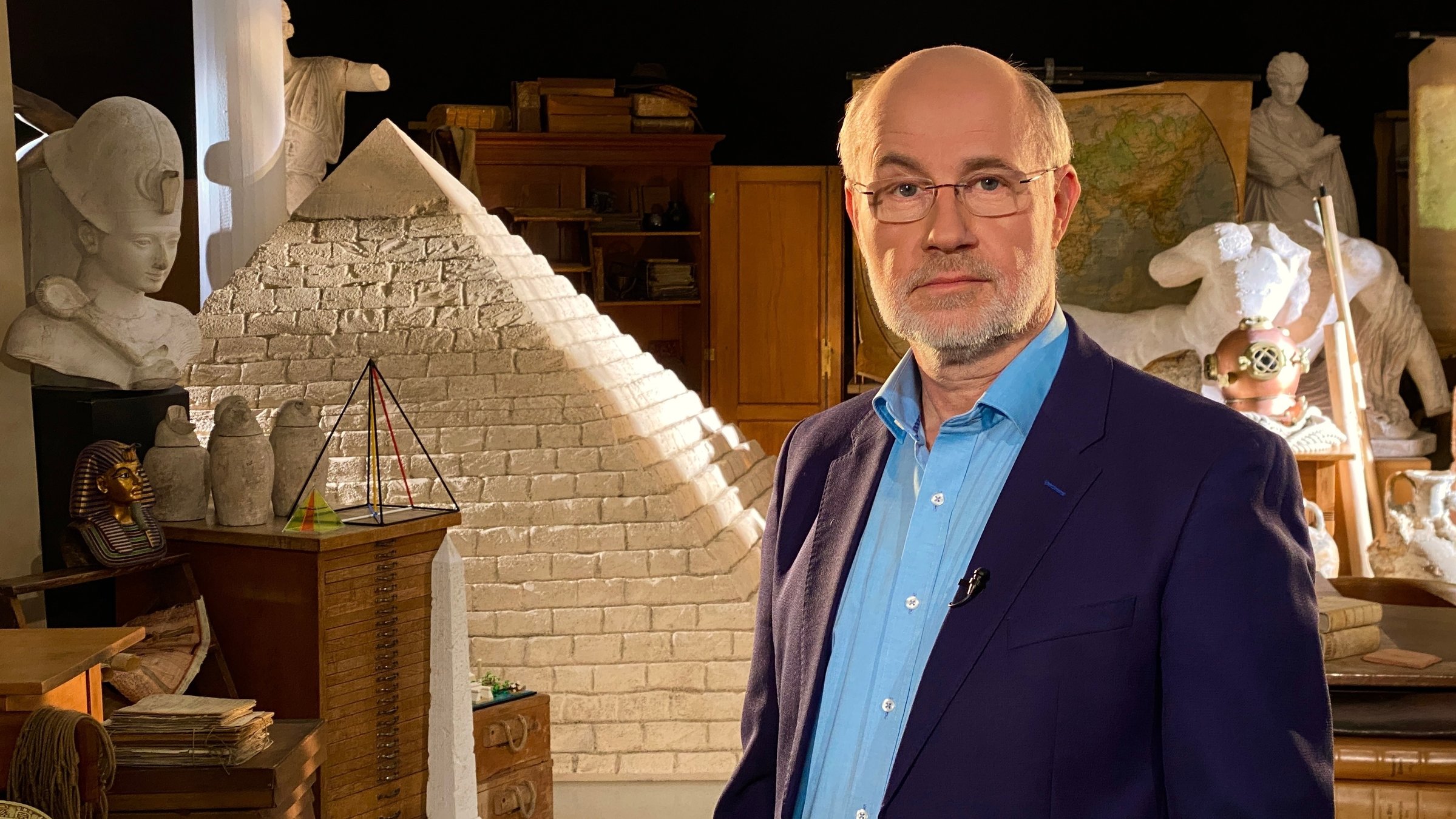 "Ungelöste Fälle der Archäologie - Pyramiden": Der Moderator Harald Lesch in einem Raum mit Nachbildungen diverser Artefakte aus der Antike, darunter ein großes Modell einer Pyramide.