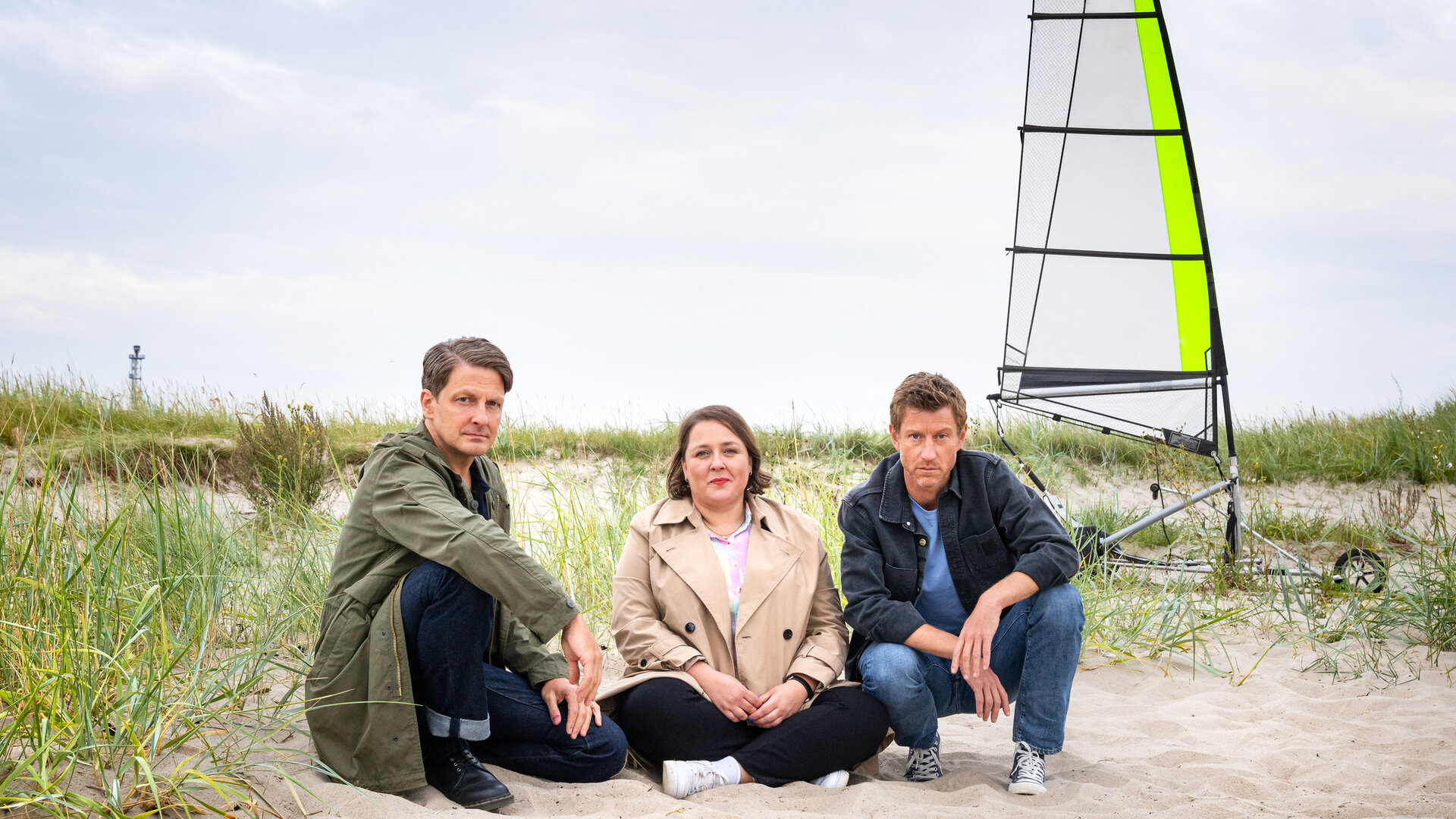 "Friesland - Geisterstunde": Wolfgang Habedank (Holger Stockhaus), Melanie Harms (Tina Pfurr) und Henk Cassens (Maxim Mehmet) sitzen im Sand einer Dünenlandschaft, im Hintergrund steht ein Strandsegler.