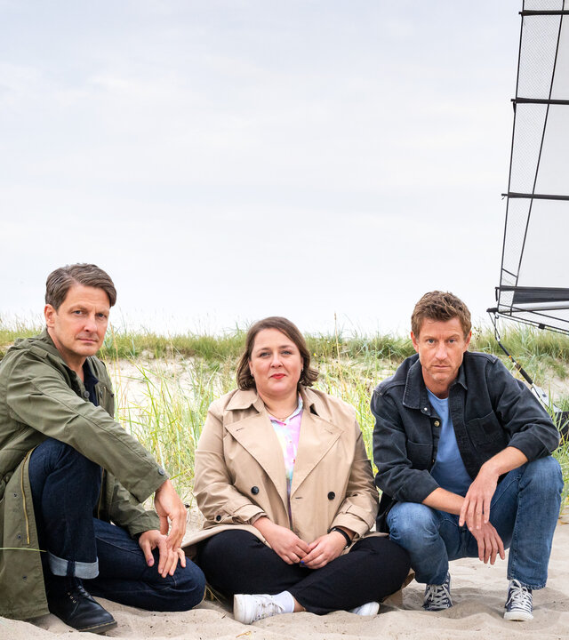 "Friesland - Geisterstunde": Wolfgang Habedank (Holger Stockhaus), Melanie Harms (Tina Pfurr) und Henk Cassens (Maxim Mehmet) sitzen im Sand einer Dünenlandschaft, im Hintergrund steht ein Strandsegler.