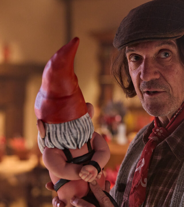 "Gnomes - Angriff der Killerzwerge - Wo ist die Königin?": Tobias Black (Syd Brisbane) steht in einem Innenraum und hält einen Gartenzwerg mit roter Mütze und grüner Schürze in den Händen. Er trägt eine Mütze, sein halblanges braunes Haar fällt darunter hervor, sein Blick geht zur Seite.