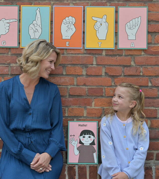"Nächste Ausfahrt Glück - Endlich ich": Katharina (Valerie Niehaus) steht im Kindergarten neben Greta (Marie Hajduk) an eine Mauer gelehnt. Die beiden schauen sich an. Zwischen und hinter ihnen hängen mehrere Ausdrucke für Gebärdensprache.