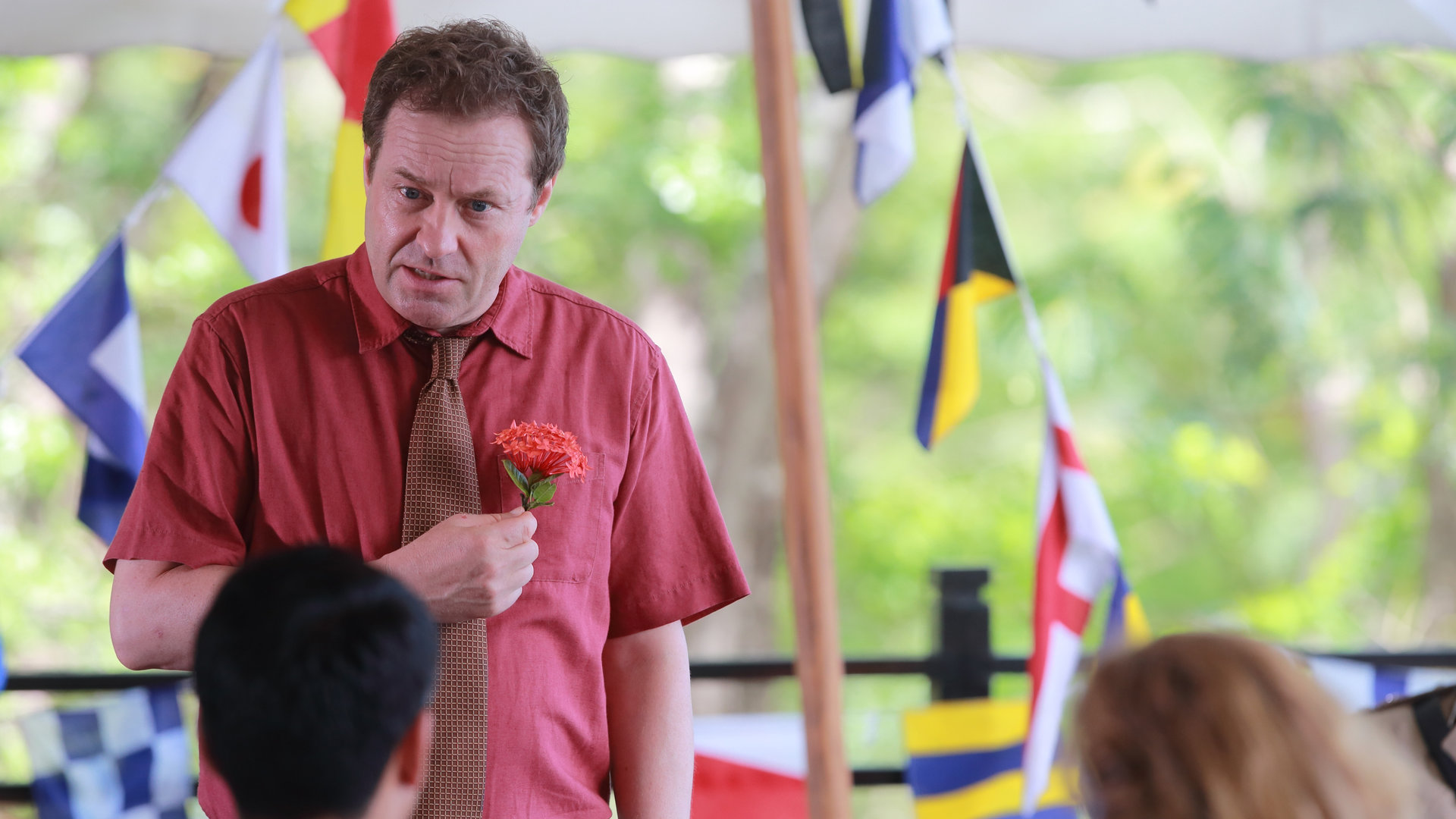 "Death in Paradise - Geldgier": DI Jack Mooney (Ardal O''Hanlon) hält eine Blume in der Hand, im Hintergrund sind verschiedene Länderflaggen zu erkennen.