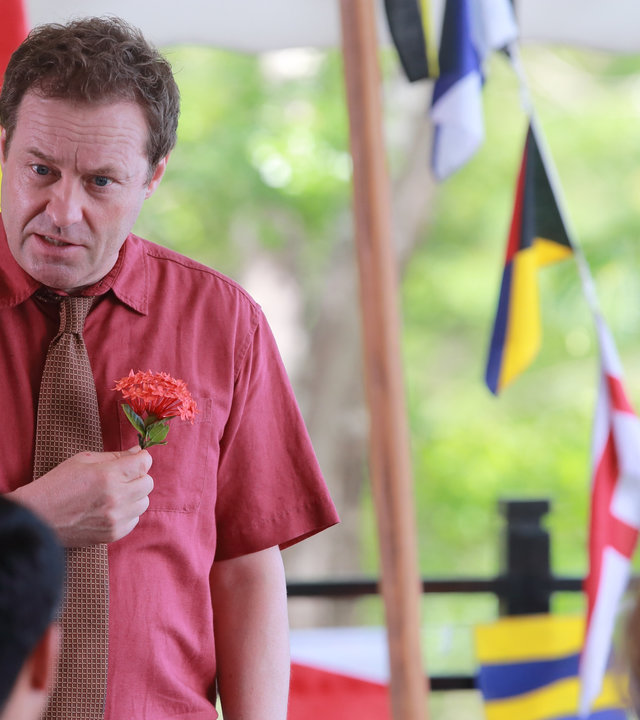 "Death in Paradise - Geldgier": DI Jack Mooney (Ardal O''Hanlon) hält eine Blume in der Hand, im Hintergrund sind verschiedene Länderflaggen zu erkennen.