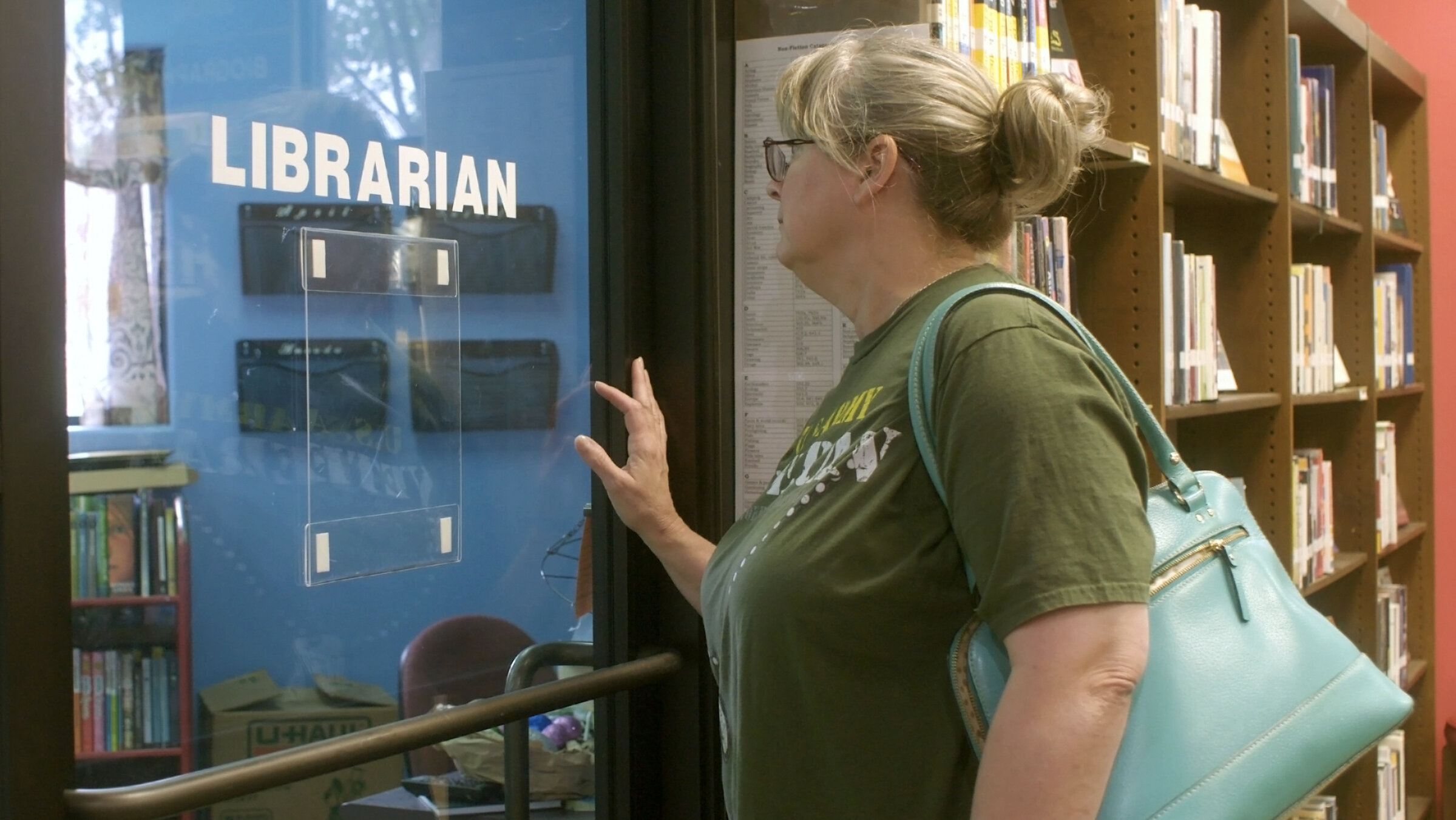 „Verbotene Bücher - Kulturkampf in den USA": Frau mit Hochsteckfrisur und türkisfarbener Handtasche steht vor einer Glastür mit dem Schild 'LIBRARIAN' in einer Bibliothek.