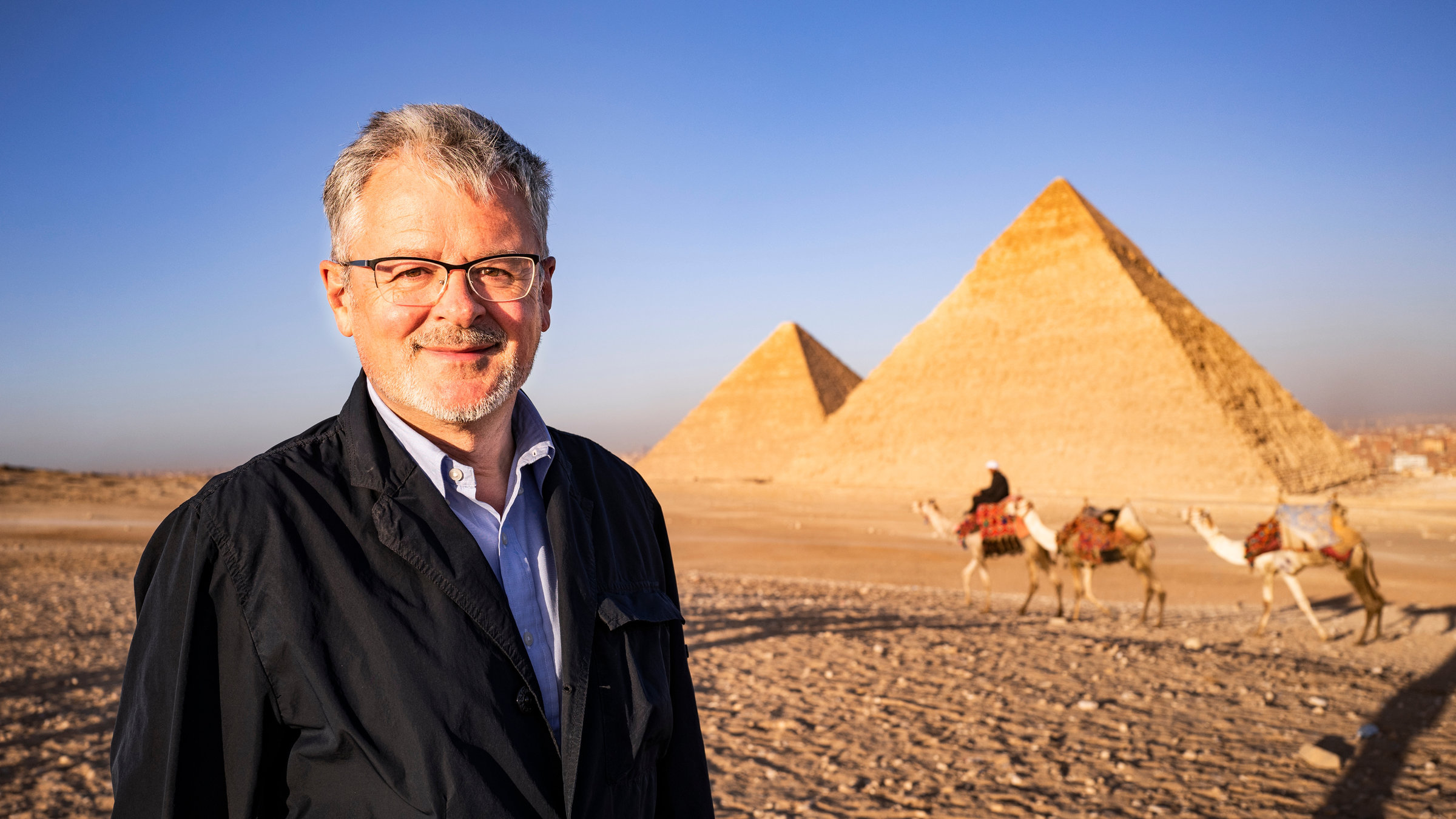 "Terra X: Welten-Saga - Die Schätze Afrikas": Außenaufnahme von den Pyramiden von Gizeh. Moderator Christopher Clark lächelt in die Kamera. Im Hintergrund ein Mann mit drei Dromedaren.