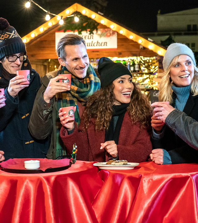 "SOKO Leipzig - Das Weihnachtswunder": Prof. Rossi (Anna Stieblich), Lorenz Rettig (Daniel Steiner), Moritz Brenner (Johannes Hendrik Langer), Kim Nowak (Amy Mußul), Ina Zimmermann (Melanie Marschke) und Jan Maybach (Marco Girnth) sind auf dem Weihnachtsmarkt. Sie stehen vor einem Glühweinstand um zwei Tische herum und stoßen an. Draußen ist es schon dunkel.