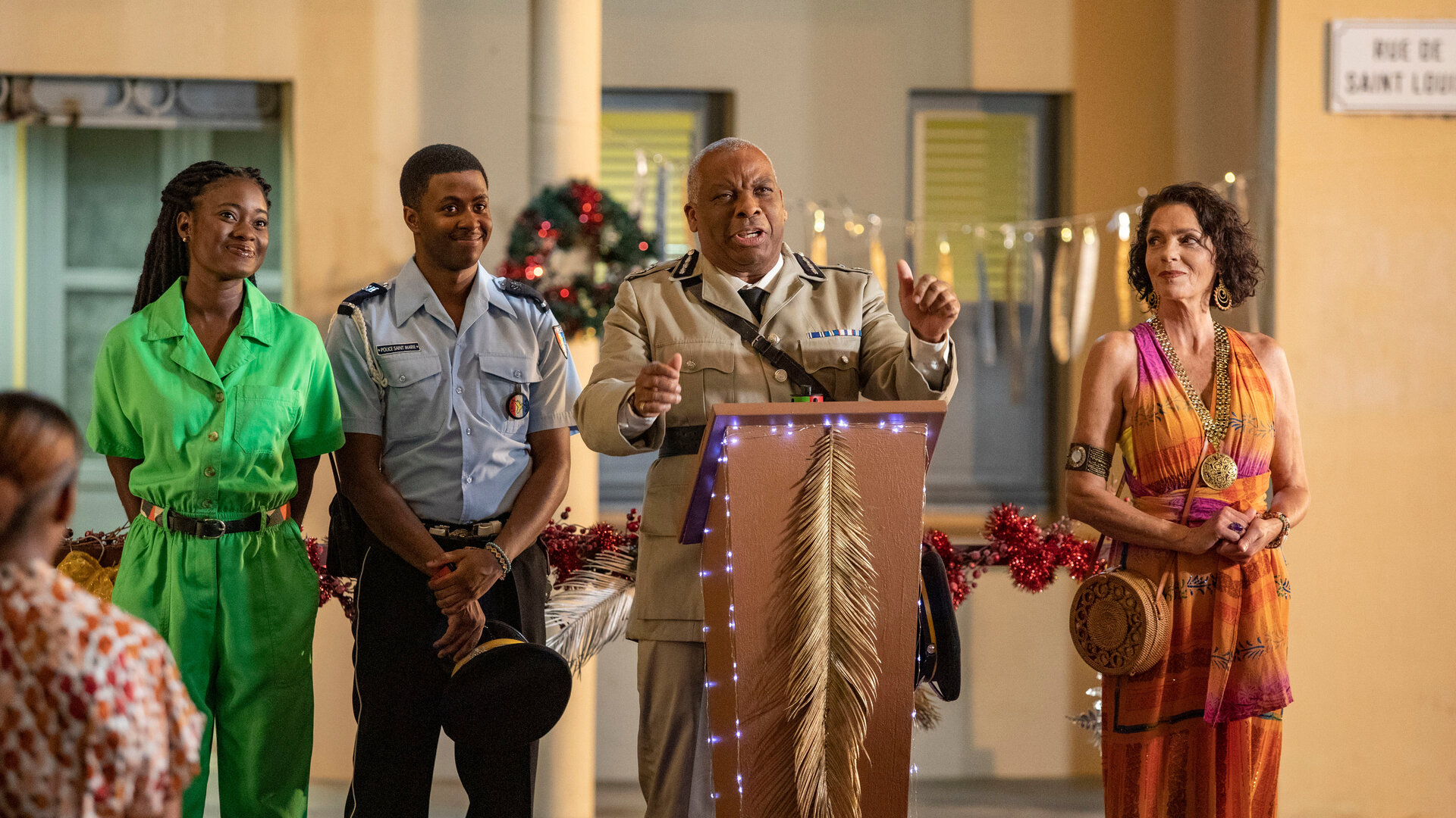 "Death in Paradise - Die letzte Botschaft (1)": Commissioner Selwyn Patterson (Don Warrington) steht an einem festlich geschmückten Rednerpult vor einem Gebäude. An seiner Seite stehen Marlon Pryce (Tahj Miles) und Naomi Thomas (Shantol Jackson), die lächelnd geradeaus blicken, sowie Catherine Bordey (Elizabeth Bourgine), die erwartungsvoll zum Commissioner schaut.