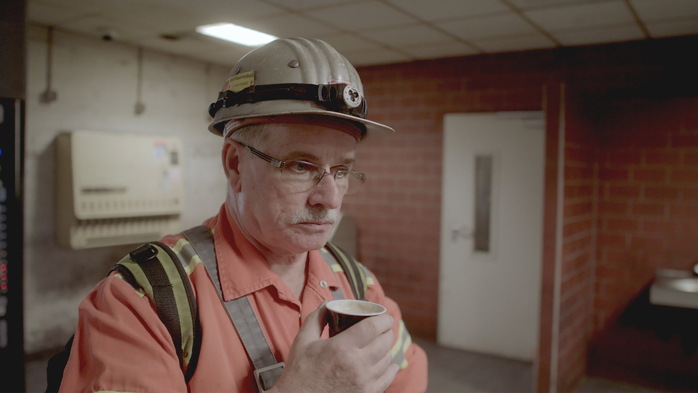 "Nach der Arbeit": Mann in Arbeitskleidung und Schutzhelm, der einen Becher mit Automatenkaffee in der Hand hält.