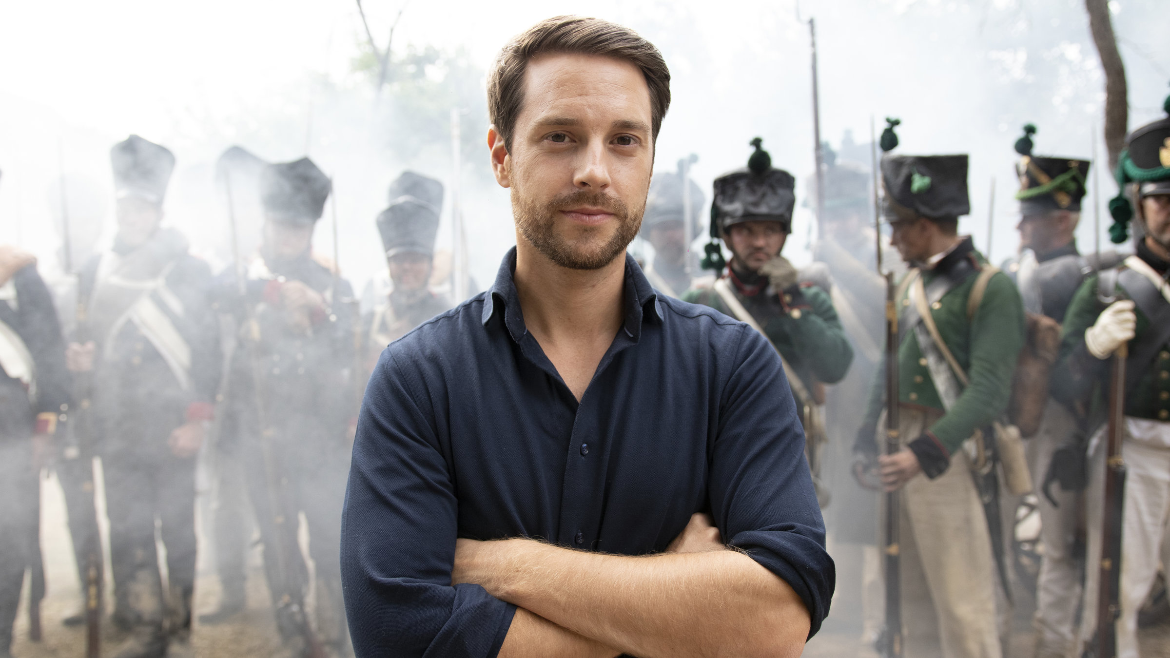 "Terra X: Ein Moment in der Geschichte - Die Völkerschlacht bei Leipzig": Moderator Mirko Drotschmann steht mit verschränkten Armen vor einer Gruppe von Darstellern in Uniformen.