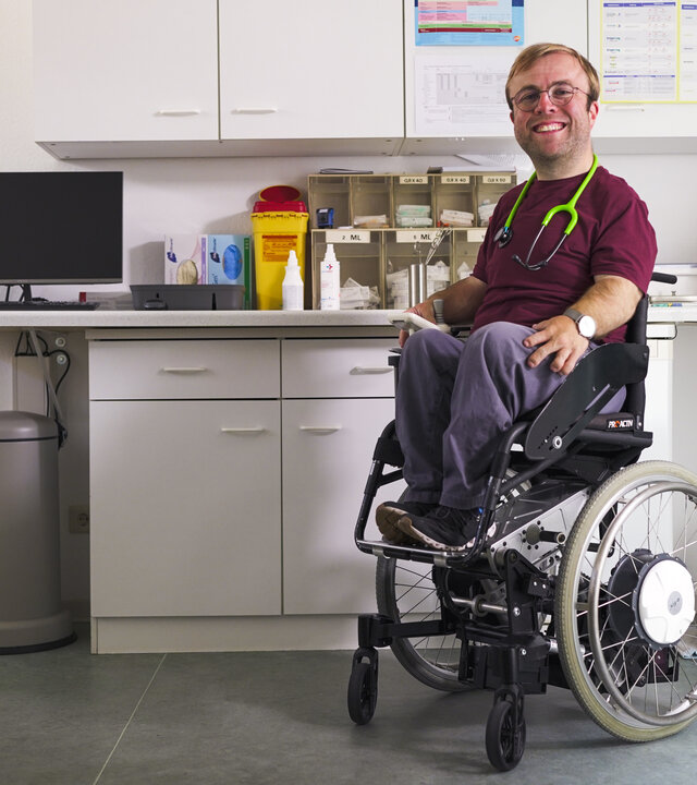 "einfach Mensch - Leopold Rupp: Ich bin Arzt": Dr. Leopold Rupp sitzt in einem elektrischen Rollstuhl vor einem Arzneischrank. Er hat ein grünes Stethoskop um den Hals hängen und lacht in die Kamera.