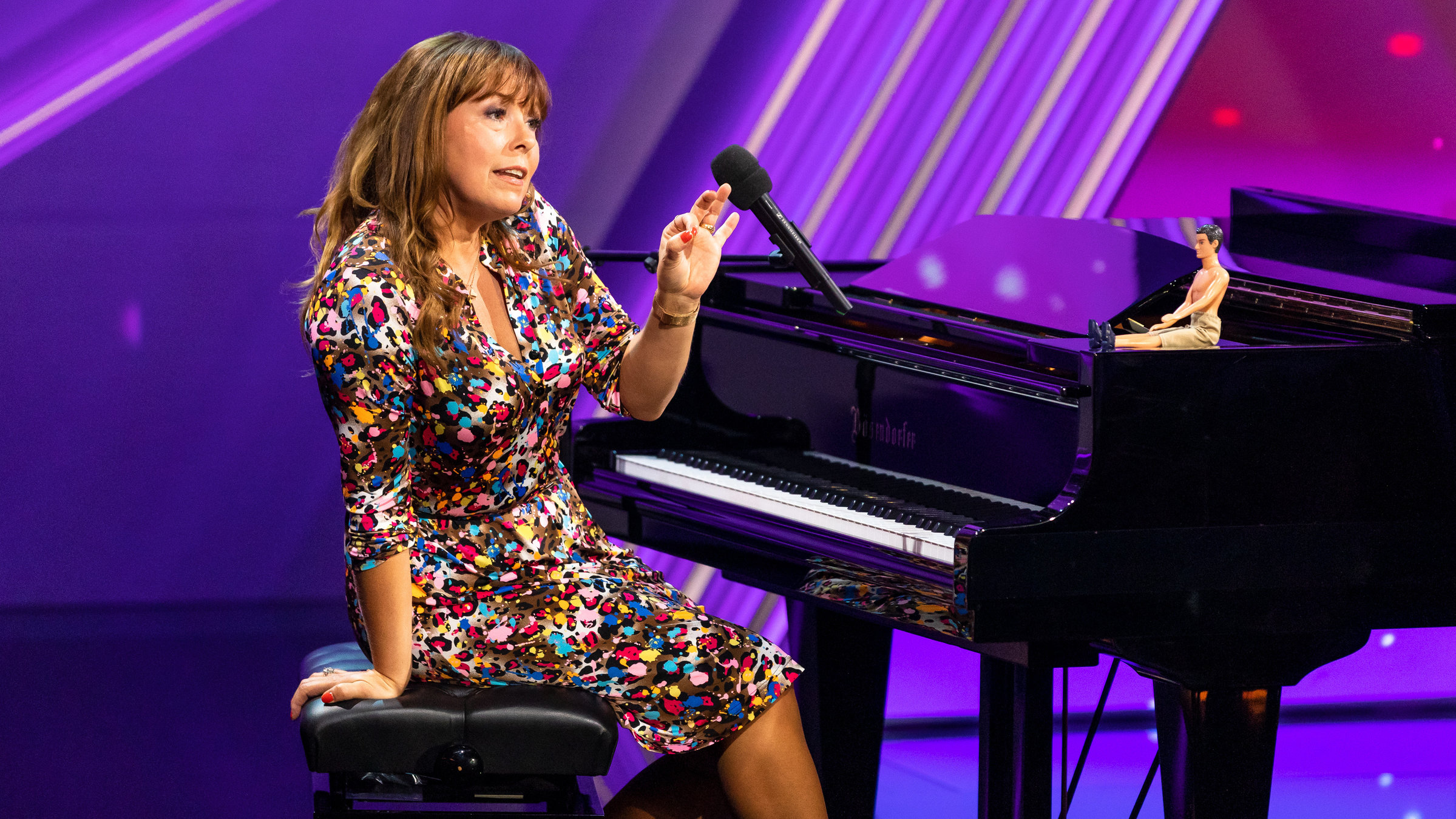"Katie Freudenschuss: Einfach Compli-Katie!": Katie Freudenschuss sitzt in einem bunten Kleid auf der 3satfestival-Bühne an einem schwarzen Flügel und gestikuliert. Auf dem Rand des Flügels sitzt eine kleine, männchliche Puppe mit freien Oberkörper.