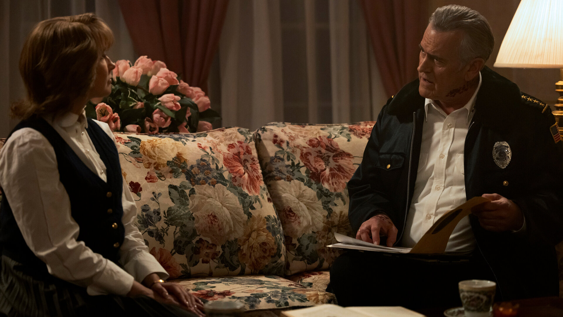 "Hysteria! - Mit Vollgas in die Hölle": Im Wohnzimmer: Tracy (Anna Camp) sitzt auf einem Sofa mit floralem Muster, während Chief Dandridge (Bruce Campbell) mit einer Akte in der Hand neben ihr sitzt und sie mit ernstem Blick ansieht. Hinter dem Sofa steht ein rosa Blumenstrauß, an der Seite eine helle Tischleuchte.