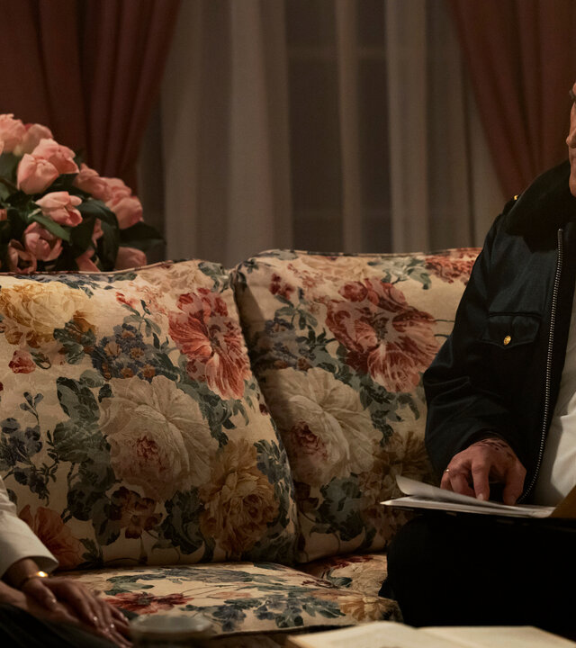 "Hysteria! - Mit Vollgas in die Hölle": Im Wohnzimmer: Tracy (Anna Camp) sitzt auf einem Sofa mit floralem Muster, während Chief Dandridge (Bruce Campbell) mit einer Akte in der Hand neben ihr sitzt und sie mit ernstem Blick ansieht. Hinter dem Sofa steht ein rosa Blumenstrauß, an der Seite eine helle Tischleuchte.