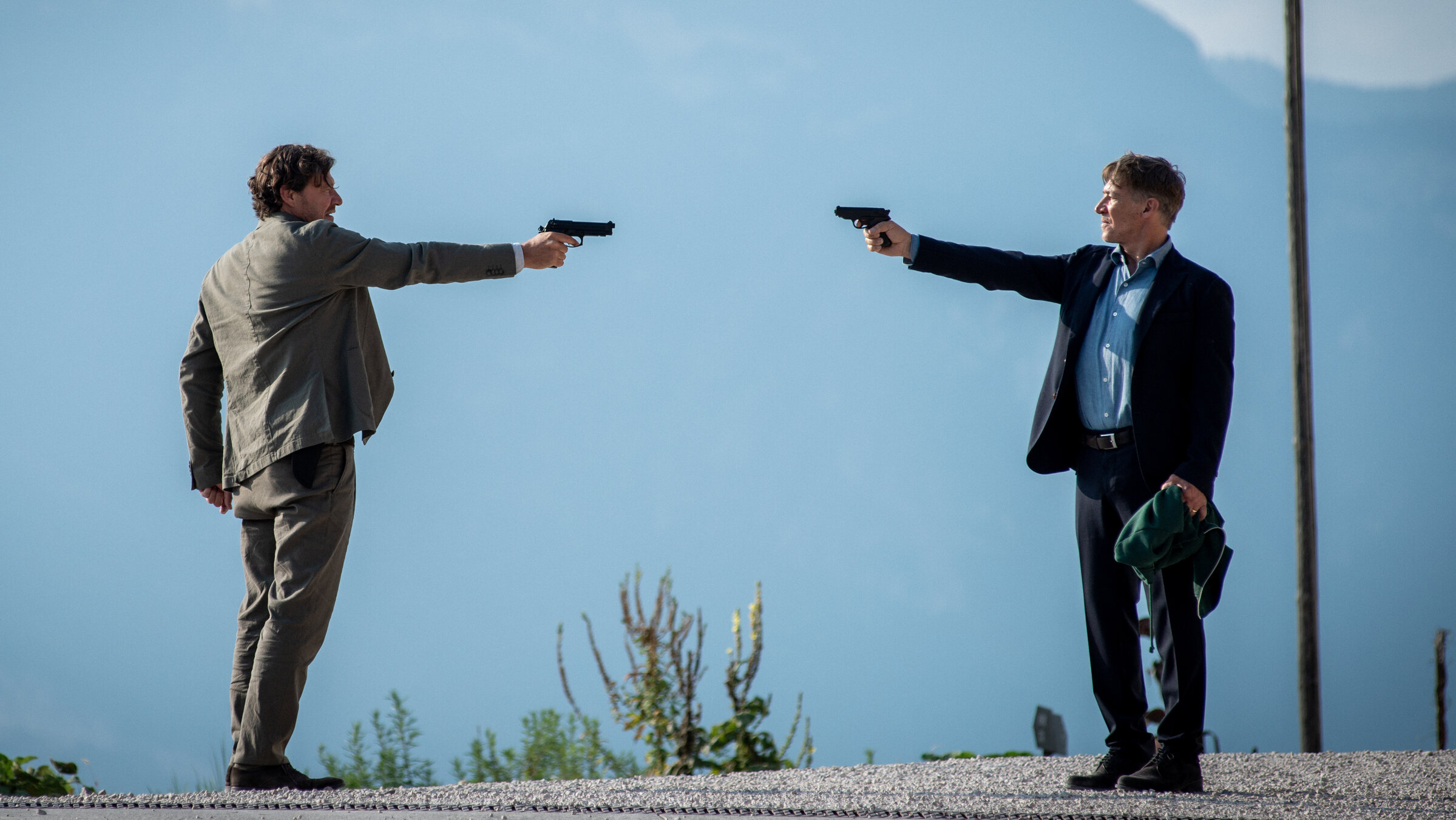 "Der Gejagte - Im Netz der Camorra": Adrin Erlacher (Harald Windisch) und Matteo DeCanin (Tobias Moretti) stehen sich auf einem Hügel gegenüber. Beide haben eine Pistole auf den jeweils anderen gerichtet, DeCanin hat in seiner anderen Hand einen grünen Pullover. Im Hintergrund lassen sich Berge erahnen.