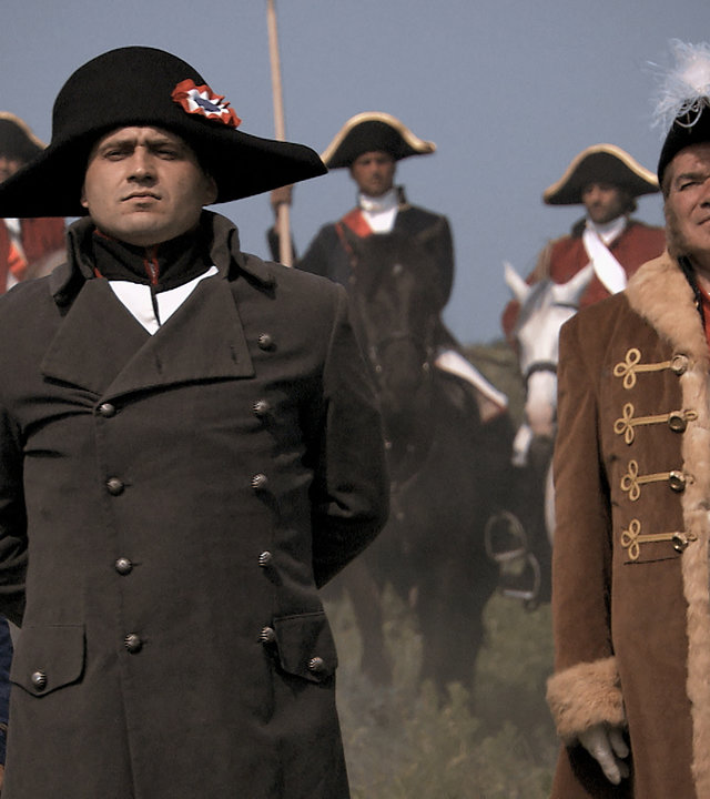 "Die glorreichen 10 - Die krassesten Raubzüge der Geschichte": Spielszene. Napoleon steht, umgeben von Offizieren, am Rande eines Schlachtfelds und schaut mit hinter dem Rücken verschränkten Armen, geschwelter Brust und erhobenen Kinn Richtung Kamera.