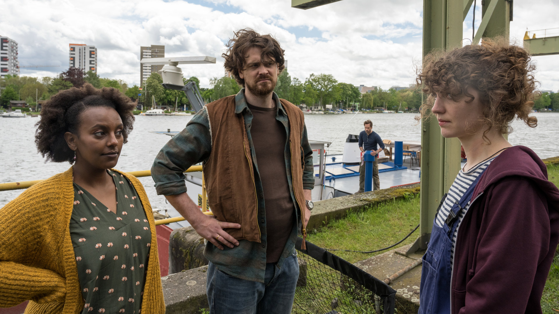 "Ein Fall für zwei - Tod auf der Zora": Sabine Pfeiffer (Yodit Tarikwa), Hanno Pfeiffer (Sebastian Fräsdorf) und Noraly van de Wal (Anne Rietmeijer) stehen am Hafen und unterhalten sich. Im Hintergrund beobachtet Leo Oswald (Wanja Mues) die Gruppe, während er an Deck der "Zora" arbeitet.