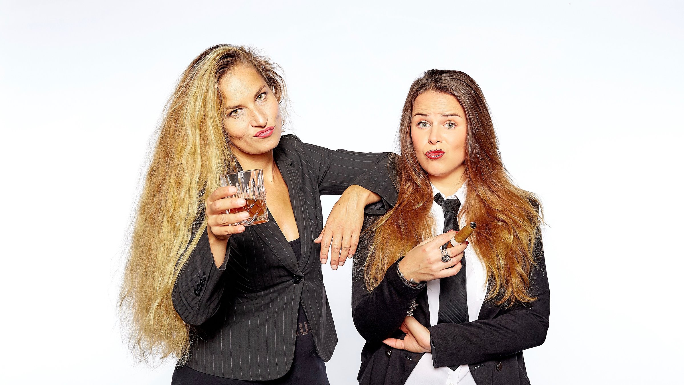 "Suchtpotenzial: Sexuelle Belustigung": Beide Frauen des Duos Suchtpotenzial stehen vor einem weißen Hintergrund dicht nebeneinander, Ariane Müller trägt eine schwärze Hose, ein schwarzes Top und einen schwarz-gestreiften Blazer, sie hält ein Whiskyglas hoch, Julia Gàmez Martin trägt eine schwarze Hose, ein weißes Hemd mit schwarzer Krawatte, dazu einen schwarzen Blazer und hält eine dicke Zigarre zwischen ihren Fingern. Beide schauen sehr selbstbewusst mit hochgezogenen Augenbrauen in die Kamera.