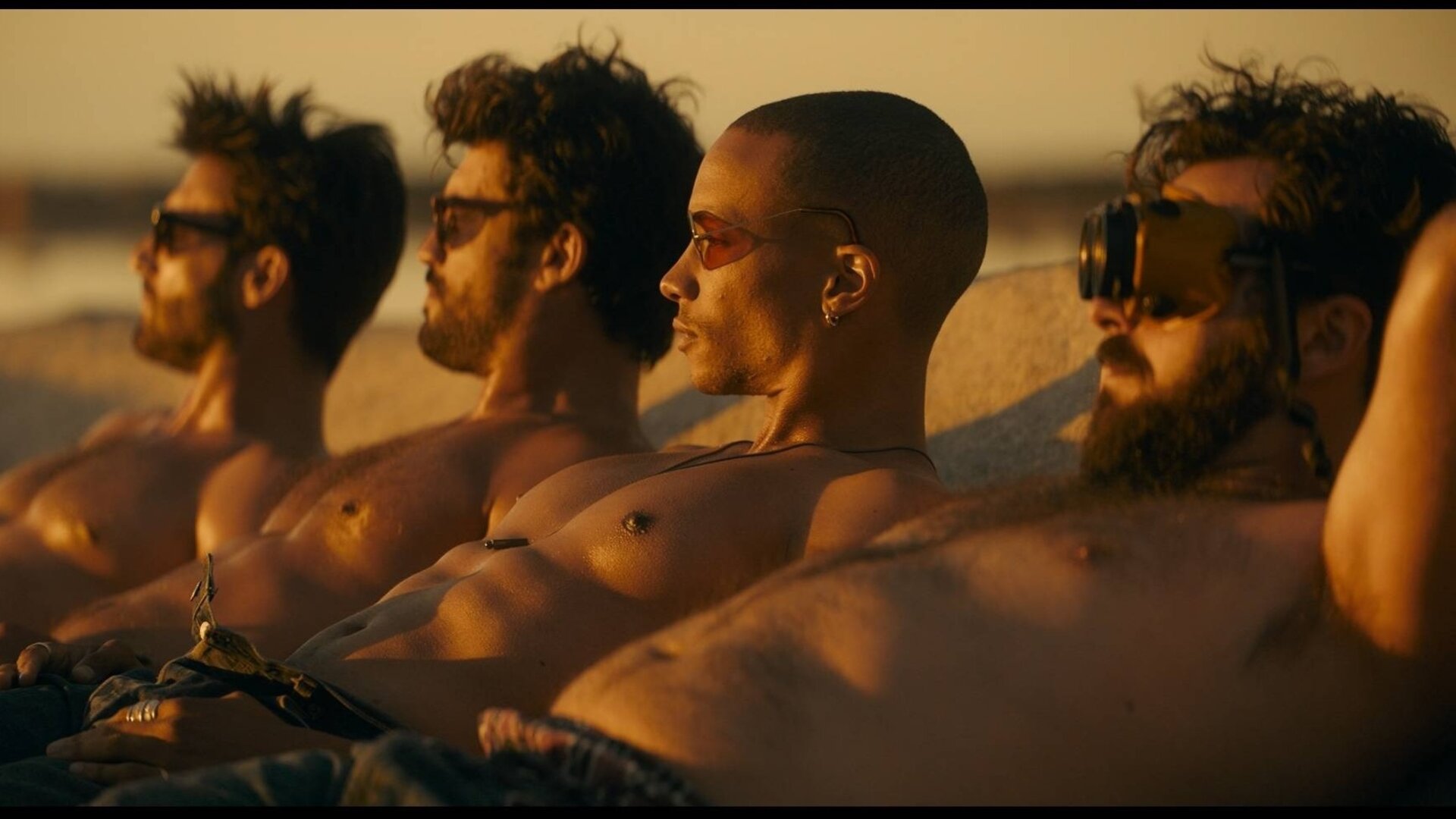 "Escort Boys - Unter Alpha-Frauen": Ben (Guillaume Labbé), Mathias (Simon Ehrlacher), Zak (Corentin Fila) und Ludo (Thibaut Evrard) liegen oberkörperfrei nebeneinander am Strand und sonnen sich. Alle tragen unterschiedliche Sonnenbrillen -  nur Ludo fällt mit einer auffälligen Schweißerbrille auf. Das Sonnenlicht blendet sie leicht, während sie in dieselbe Richtung blicken.