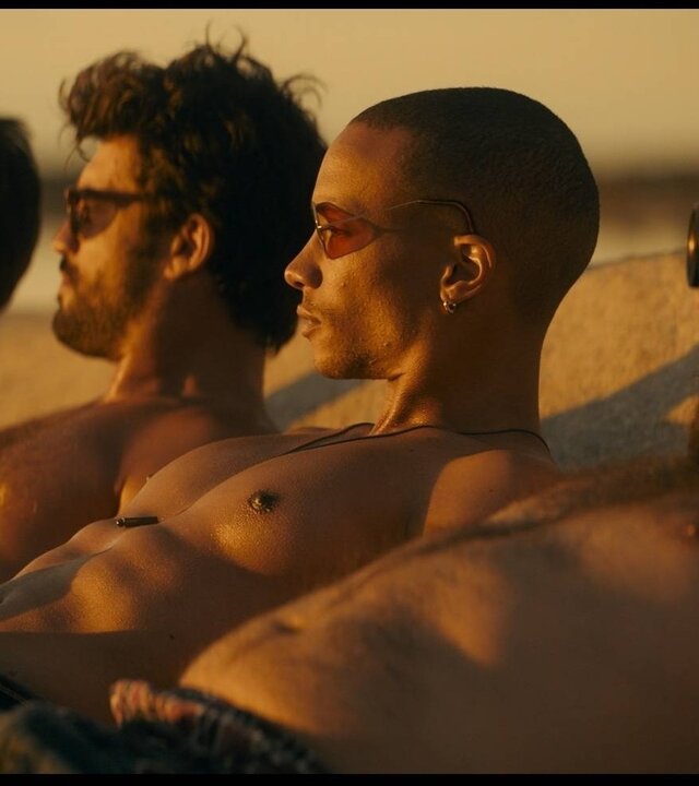 "Escort Boys - Unter Alpha-Frauen": Ben (Guillaume Labbé), Mathias (Simon Ehrlacher), Zak (Corentin Fila) und Ludo (Thibaut Evrard) liegen oberkörperfrei nebeneinander am Strand und sonnen sich. Alle tragen unterschiedliche Sonnenbrillen -  nur Ludo fällt mit einer auffälligen Schweißerbrille auf. Das Sonnenlicht blendet sie leicht, während sie in dieselbe Richtung blicken.