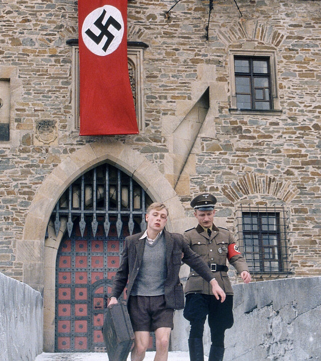 "Napola - Elite für den Führer": Über einem Burgtor hängt eine wehende Hakenkreuzflagge. Davor führt eine Soldat (Komparse) Friedrich (Max Riemelt) von der Burg weg. Friedrich hat Verletzungen am Auge und trägt einen Koffer.