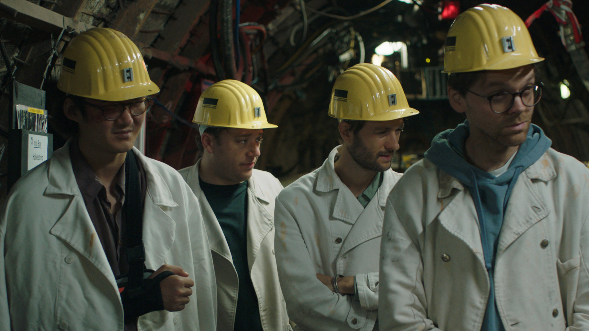 "FETT UND FETT - So kompliziert": Bulli (Bulgan Molor-Erdene), Jaksch (Jakob Schreier), Jogi (Oscar Lauterbach) und Nicolai (Nicolai Zeitler) stehen nebeneinander in einem Bergbau-Tunnel mit vielen Kabeln. Sie tragen alle weiße Jacken und gelbe Schutzhelme.