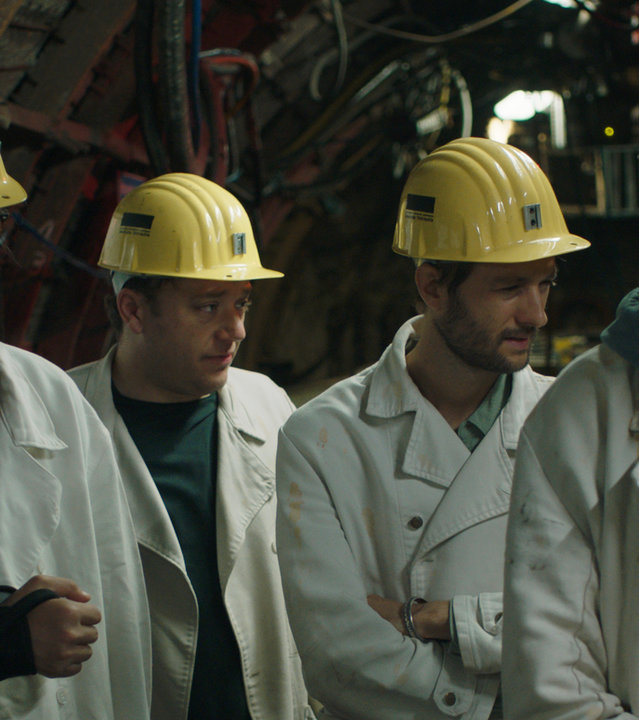 "FETT UND FETT - So kompliziert": Bulli (Bulgan Molor-Erdene), Jaksch (Jakob Schreier), Jogi (Oscar Lauterbach) und Nicolai (Nicolai Zeitler) stehen nebeneinander in einem Bergbau-Tunnel mit vielen Kabeln. Sie tragen alle weiße Jacken und gelbe Schutzhelme.
