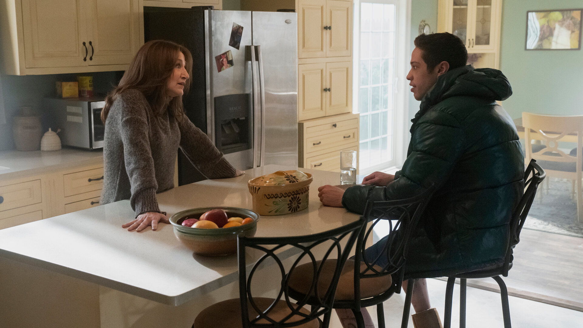 "Bupkis - Der Vergnügungspark": Amy (Edie Falco) steht an der weißen Küchentheke mit ihrem Sohn Pete (Pete Davidson). Pete sitzt ihr gegenüber in seiner dicken Daunenjacke und hört ihr zu. Die Küche ist im Landhausstil gestaltet und das Zimmer ist vom Tageslicht durchströmt. Am Kühlschrank kleben Fotos.