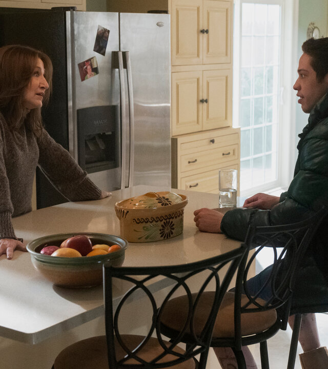 "Bupkis - Der Vergnügungspark": Amy (Edie Falco) steht an der weißen Küchentheke mit ihrem Sohn Pete (Pete Davidson). Pete sitzt ihr gegenüber in seiner dicken Daunenjacke und hört ihr zu. Die Küche ist im Landhausstil gestaltet und das Zimmer ist vom Tageslicht durchströmt. Am Kühlschrank kleben Fotos.