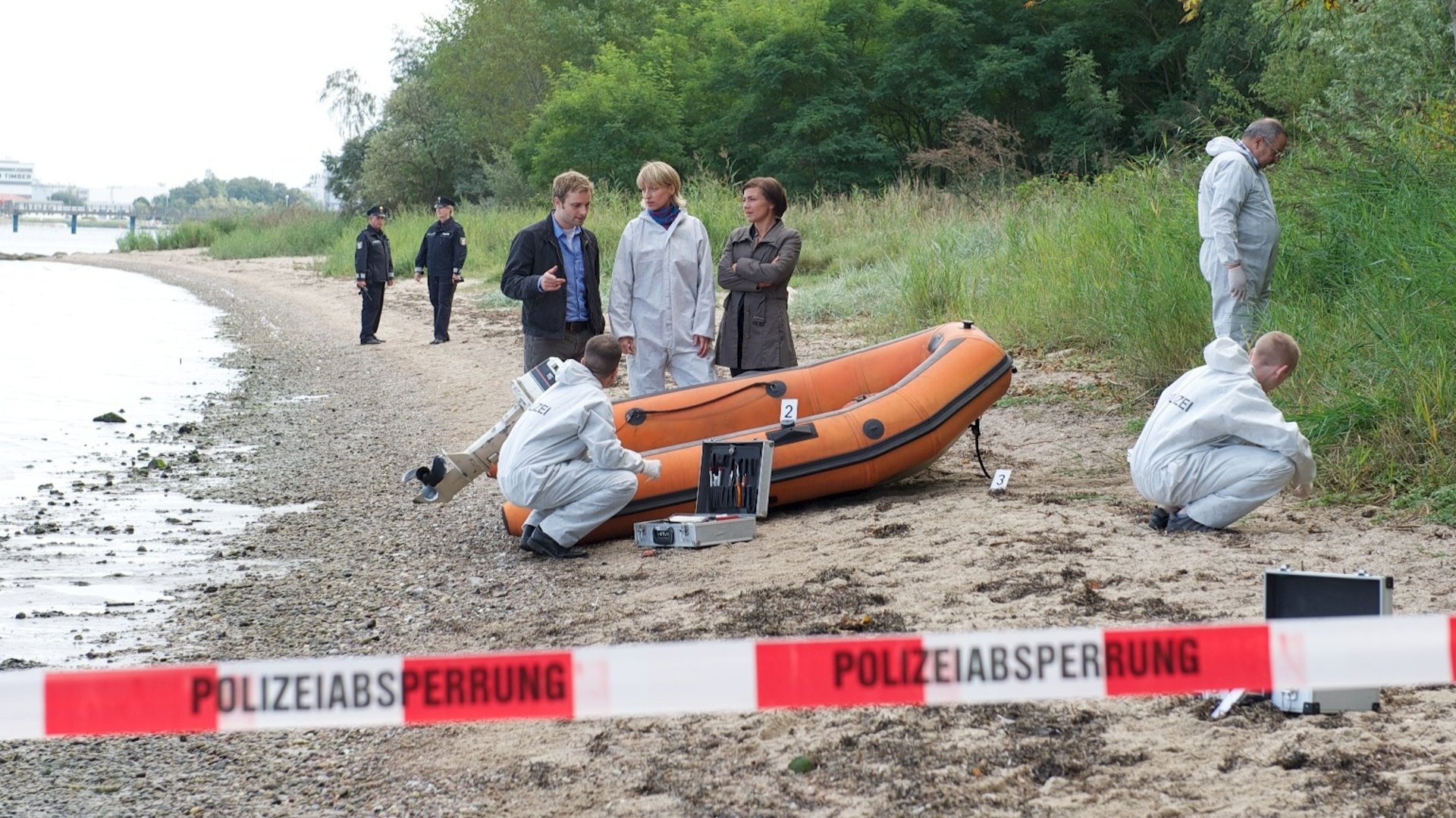 "SOKO Wismar - Tod auf See": An einem Strand liegt ein orangefarbenes Schlauchboot. Dahinter stehen Nils Theede (Jonas Laux), Roswitha Prinzler (Silke Matthias) und Katrin Börensen (Claudia Schmutzler) und besprechen sich. (Rest: Komparsen)
