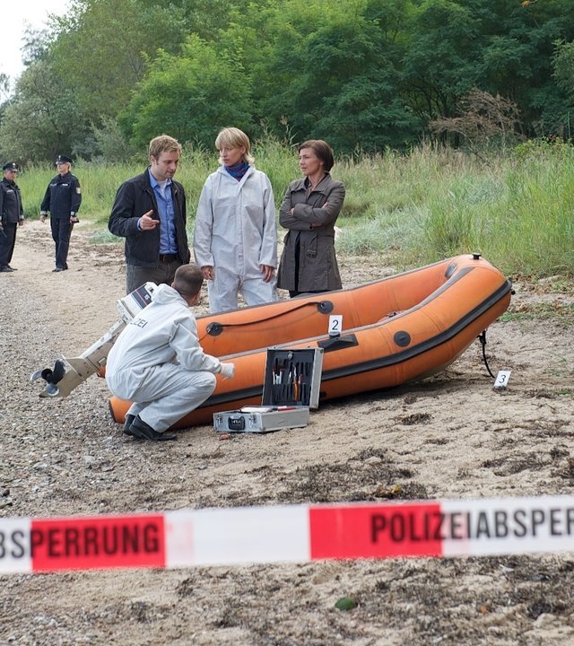"SOKO Wismar - Tod auf See": An einem Strand liegt ein orangefarbenes Schlauchboot. Dahinter stehen Nils Theede (Jonas Laux), Roswitha Prinzler (Silke Matthias) und Katrin Börensen (Claudia Schmutzler) und besprechen sich. (Rest: Komparsen)