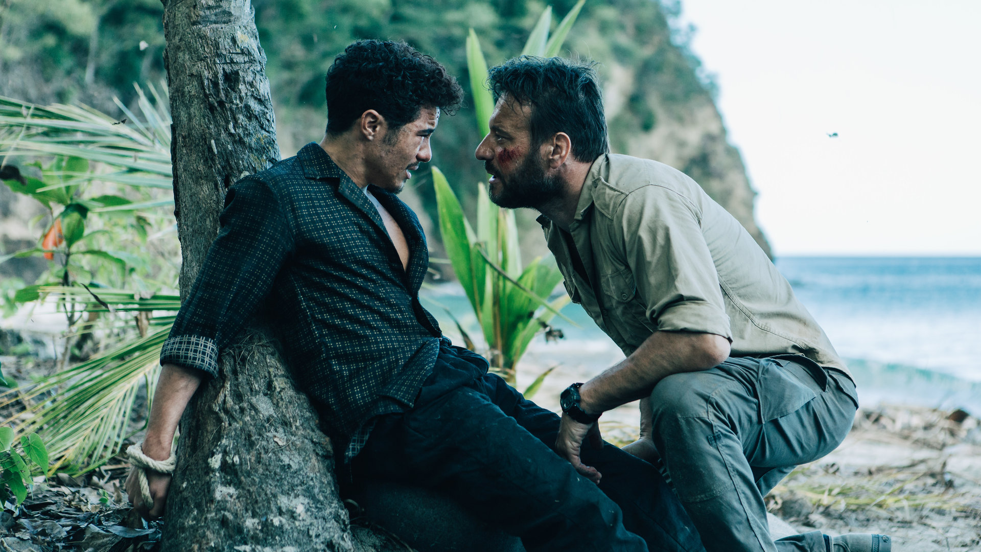 "They Were Ten": Eddy (Samy Seghir) liegt am Fuße eines Baumes am Strand, seine Hände sind hinter ihm und  dem Baum mit einem Seil gefesselt. Xavier (Samuel Le Bihan) kniet vor ihm, sein Gesicht nah an Eddys. Xavier schaut Eddy fragend und drohend an.