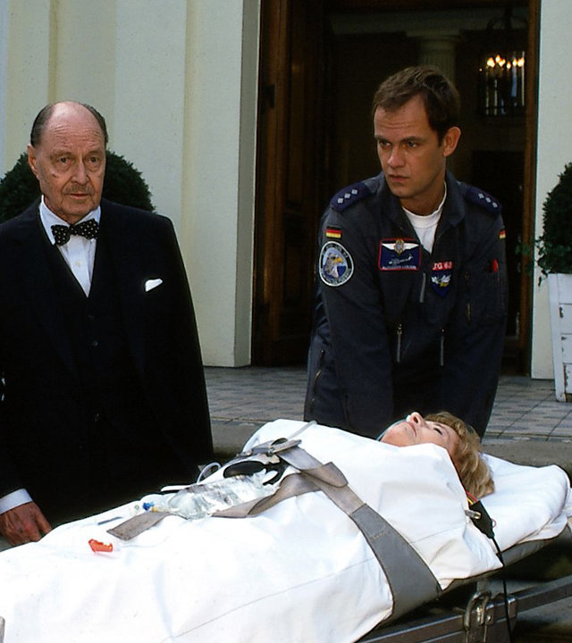 "Die Rettungsflieger - Der Neue": Die Rettungsflieger Alexander (Matthias Leja) und Torsten (Tom Wlaschiha) stehen neben der Transportliege, auf der Greta Paulsen (Gisela Uhlen) liegt. Neben ihr stehen Dietrich (Ivan Desny) und Herrmann (Heinz Dieter Knaup).