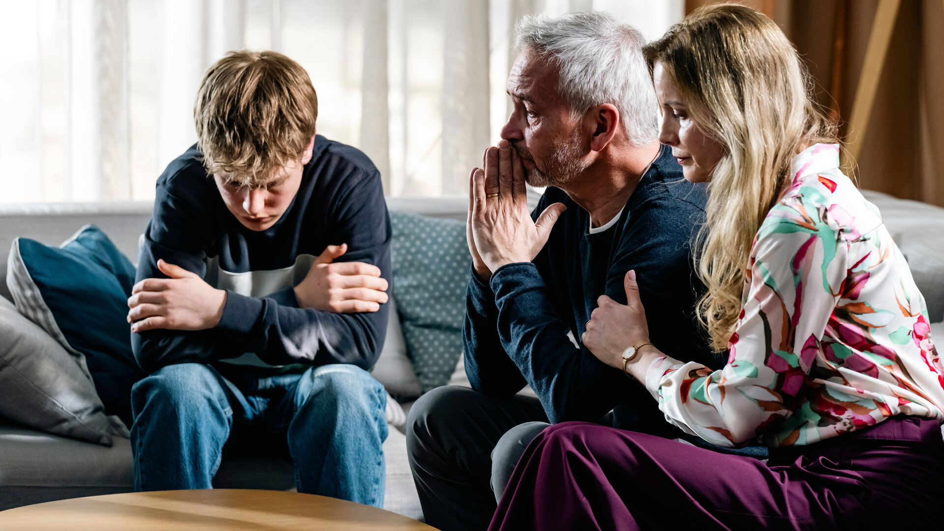 "SOKO Köln - Tödliche Rückkehr": Johannes (Thomas Dannemann), Yolanda (Pia Sarpei) und Toto Rechberg (Ted Trube) sitzen dicht nebeneinander auf einem grauen Sofa in einem hellen Raum. Toto hat die Arme vor der Brust verschränkt und beugt sich mit ernstem Gesicht nach unten. Neben ihm sitzt Johannes Rechberg, der sich mit traurigem Blick die Hände vor den Mund hält. Yolanda sitzt neben ihm und hat ihm eine Hand auf den Arm gelegt, während sie nach unten schaut.