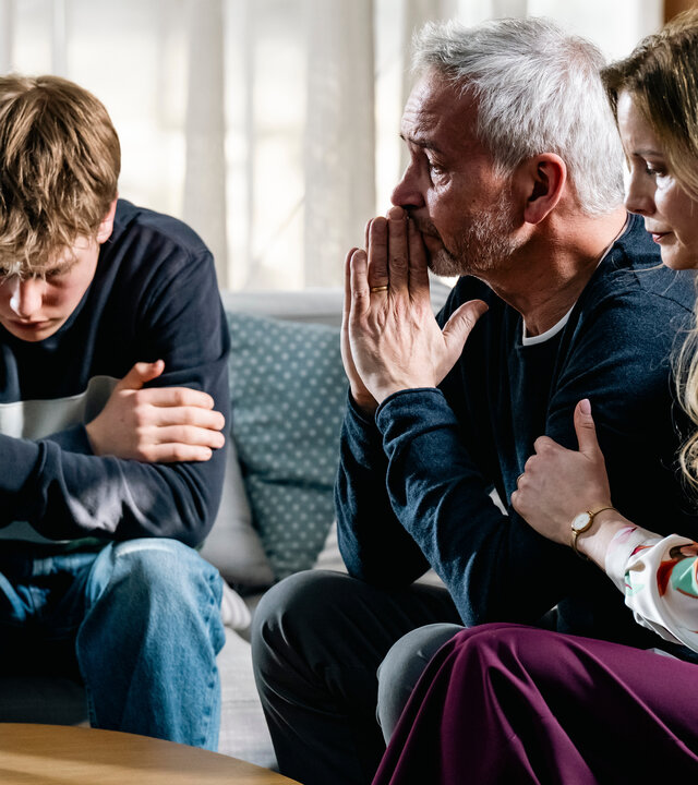 "SOKO Köln - Tödliche Rückkehr": Johannes (Thomas Dannemann), Yolanda (Pia Sarpei) und Toto Rechberg (Ted Trube) sitzen dicht nebeneinander auf einem grauen Sofa in einem hellen Raum. Toto hat die Arme vor der Brust verschränkt und beugt sich mit ernstem Gesicht nach unten. Neben ihm sitzt Johannes Rechberg, der sich mit traurigem Blick die Hände vor den Mund hält. Yolanda sitzt neben ihm und hat ihm eine Hand auf den Arm gelegt, während sie nach unten schaut.