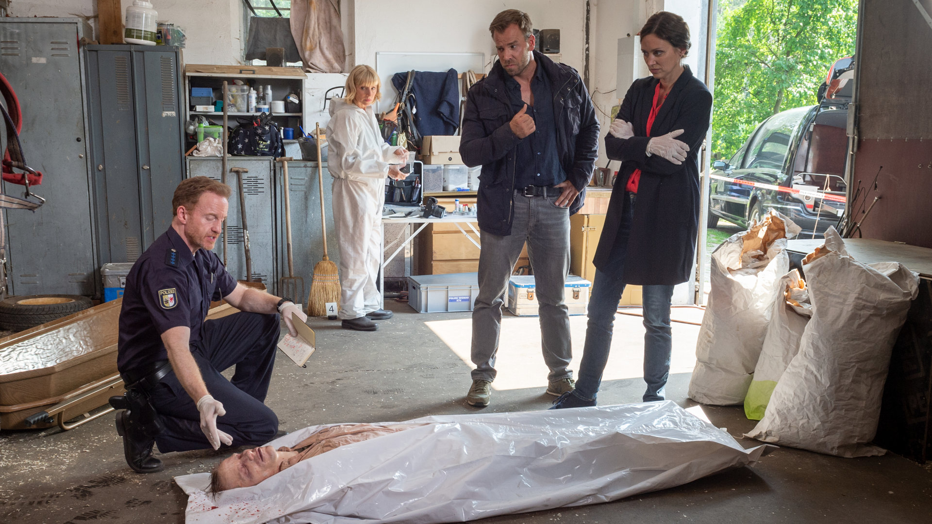 "SOKO Wismar – Ein würdeloser Tod": Im Vorraum eines Stalls liegt Erik Zierling (Alexander Petau) bereits in den Leichensack gepackt. Timmermann hockt an seinem Kopfende und klärt seine Kollegen Pöhlmann (Dominic Boeer) und Joost (Nike Fuhrmann) über die Umstände seines Todes auf. Im Hintergrund untersucht Roswitha (Silke Matthias) den Tatort.