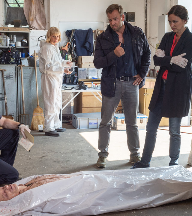 "SOKO Wismar – Ein würdeloser Tod": Im Vorraum eines Stalls liegt Erik Zierling (Alexander Petau) bereits in den Leichensack gepackt. Timmermann hockt an seinem Kopfende und klärt seine Kollegen Pöhlmann (Dominic Boeer) und Joost (Nike Fuhrmann) über die Umstände seines Todes auf. Im Hintergrund untersucht Roswitha (Silke Matthias) den Tatort.